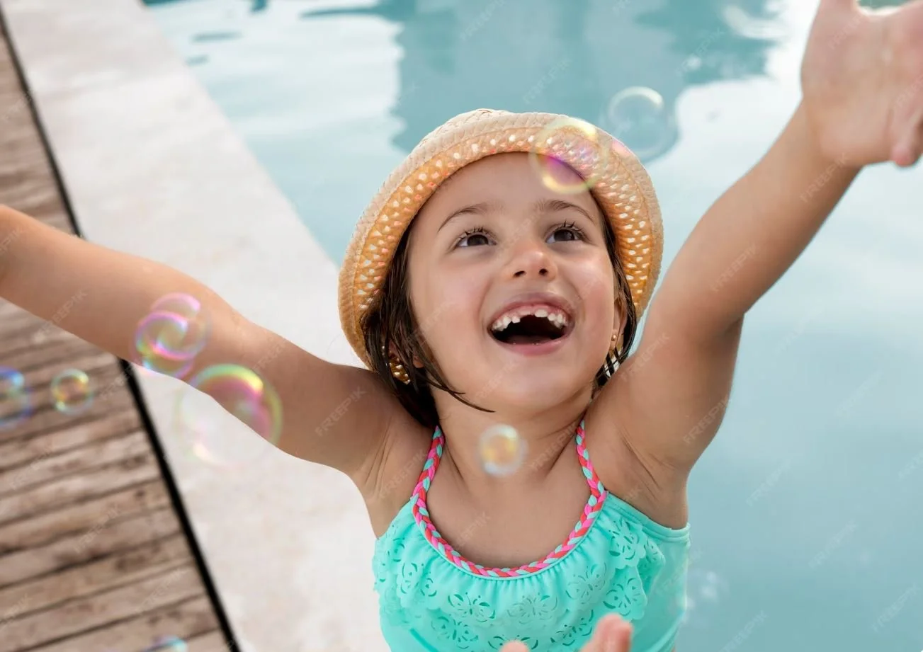 A young girl in a pool with her arms raised, wearing a straw hat and a turquoise swimsuit, smiling joyfully.