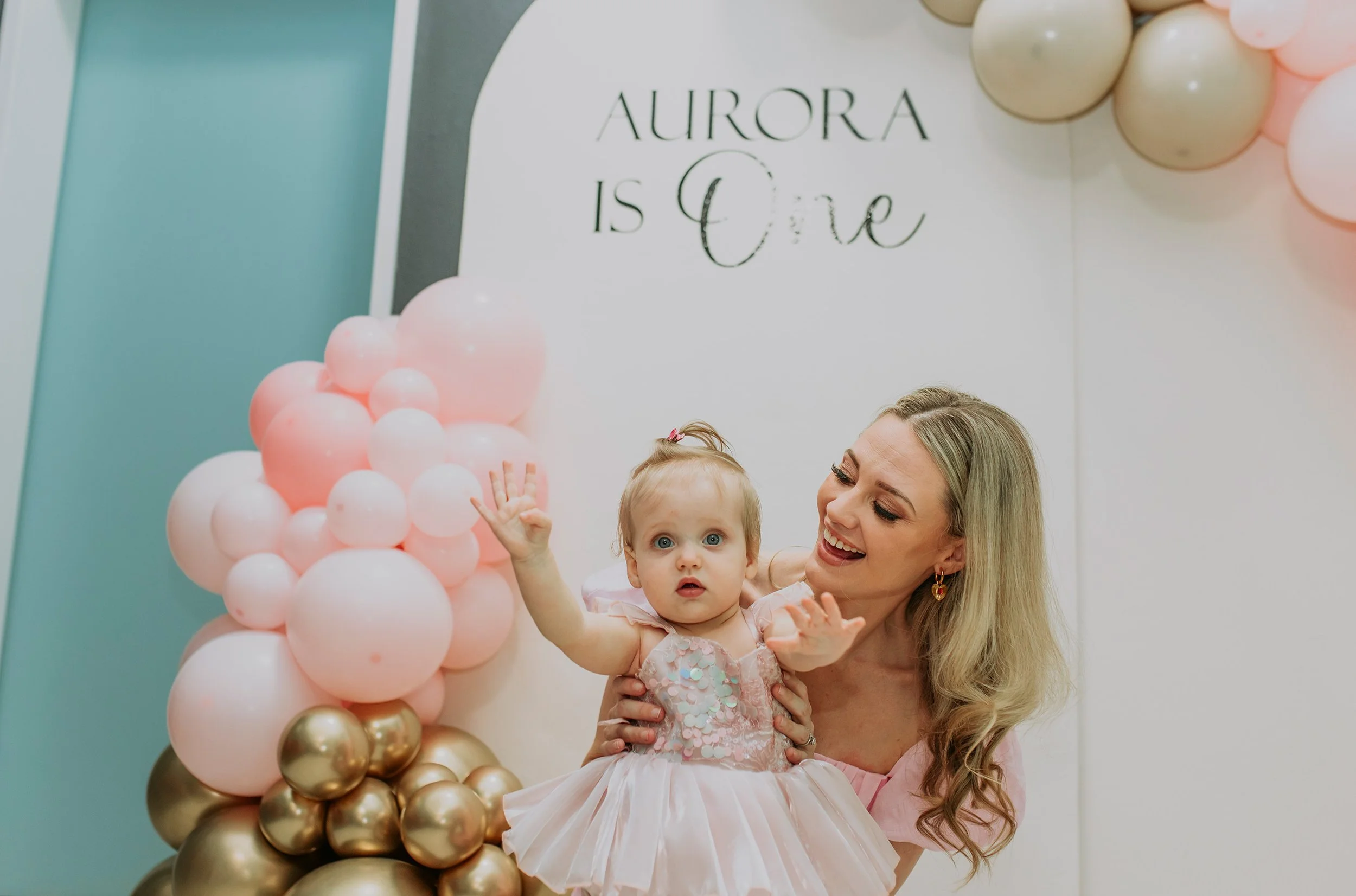 A woman and a young girl celebrating a first birthday, with balloons and a sign that reads 'Aurora is One' in the background.
