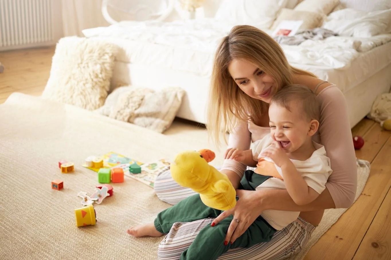 A woman and a young boy playing with a plush duck toy in a cozy, well-lit bedroom with a bed, pillows, and scattered toys.