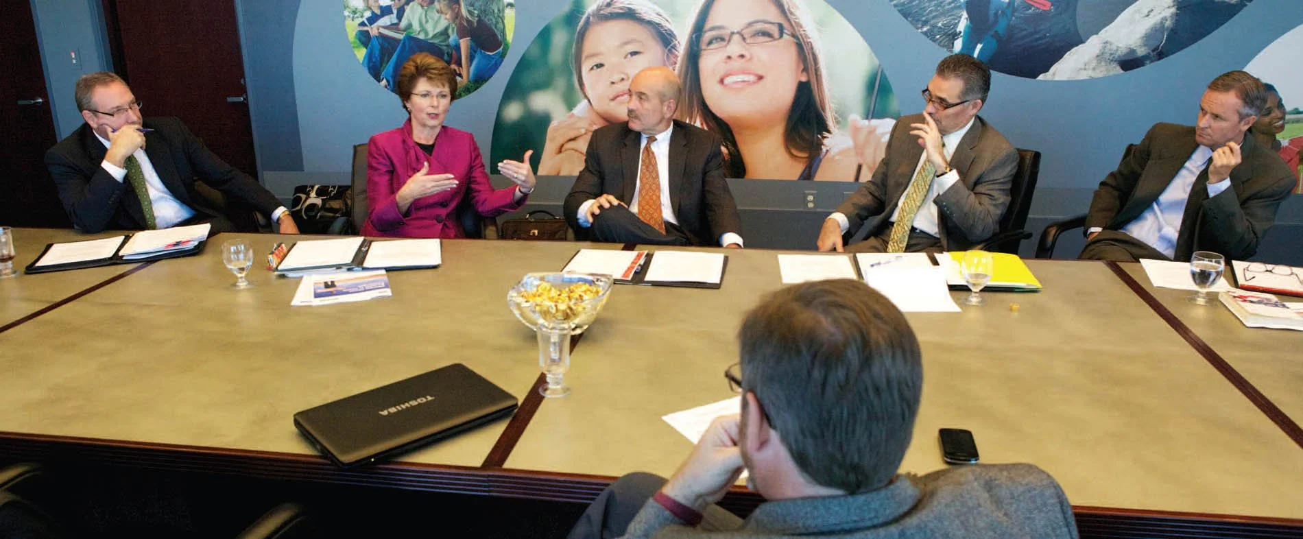Business meeting with six people seated around a conference table, with a large mural of people and nature in the background.