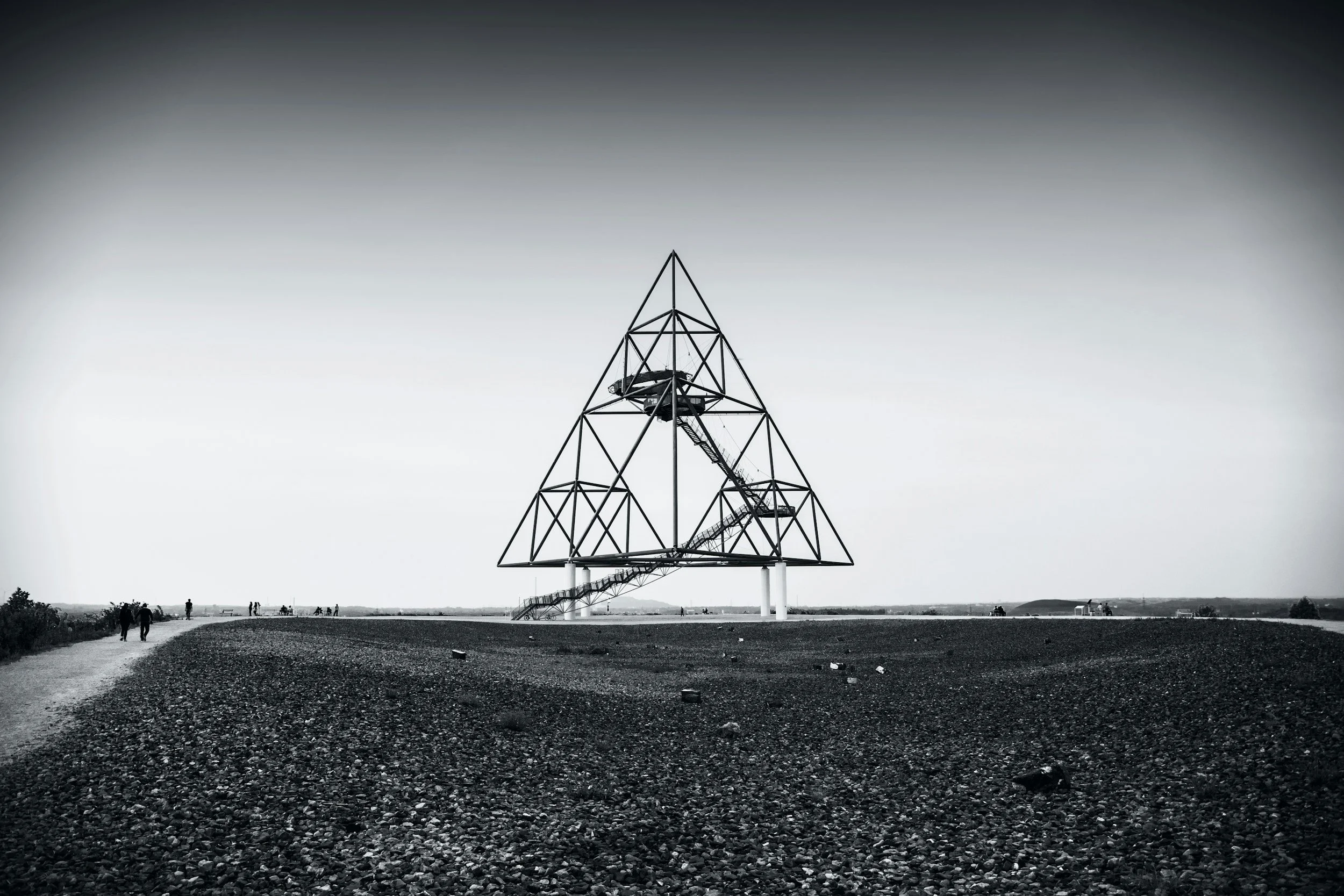 A large geometric metal structure resembling a pyramid or triangular shape, with an internal staircase, stands on a flat, gravel-covered landscape with a few small groups of people walking nearby.