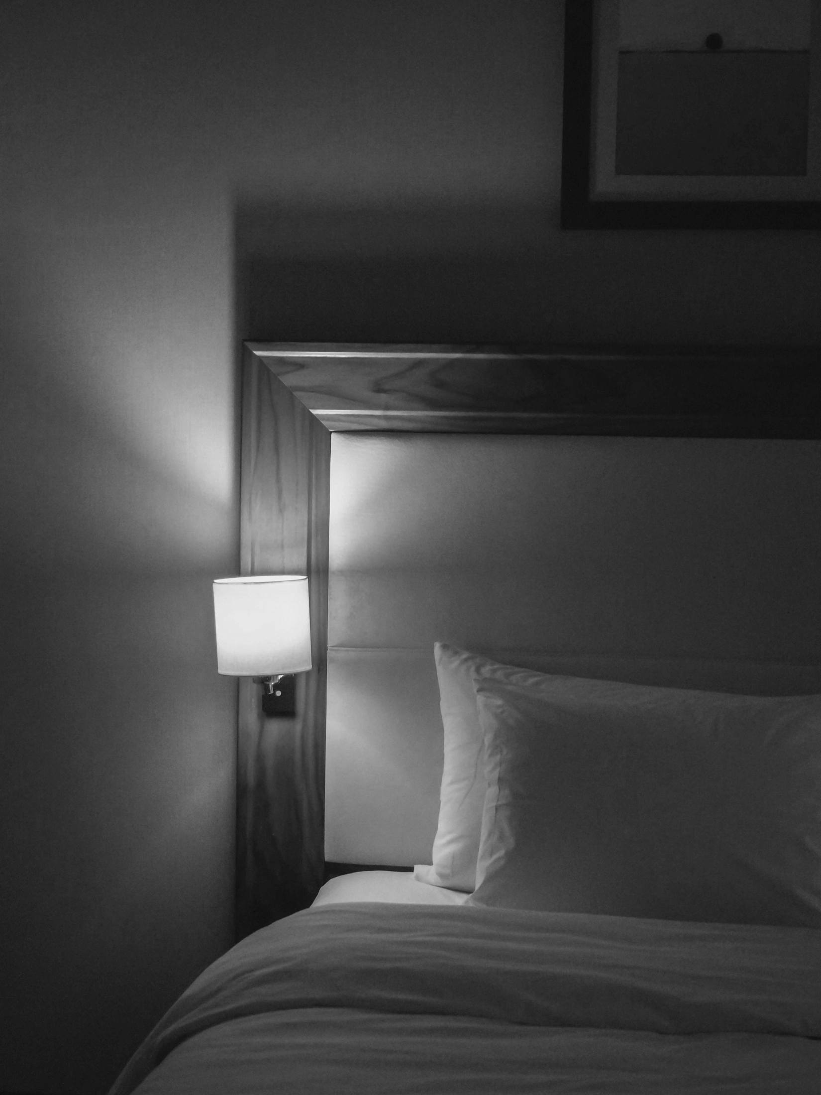 A black and white image of a bedroom with a bed, pillow, and a wall-mounted lamp next to a wooden headboard.