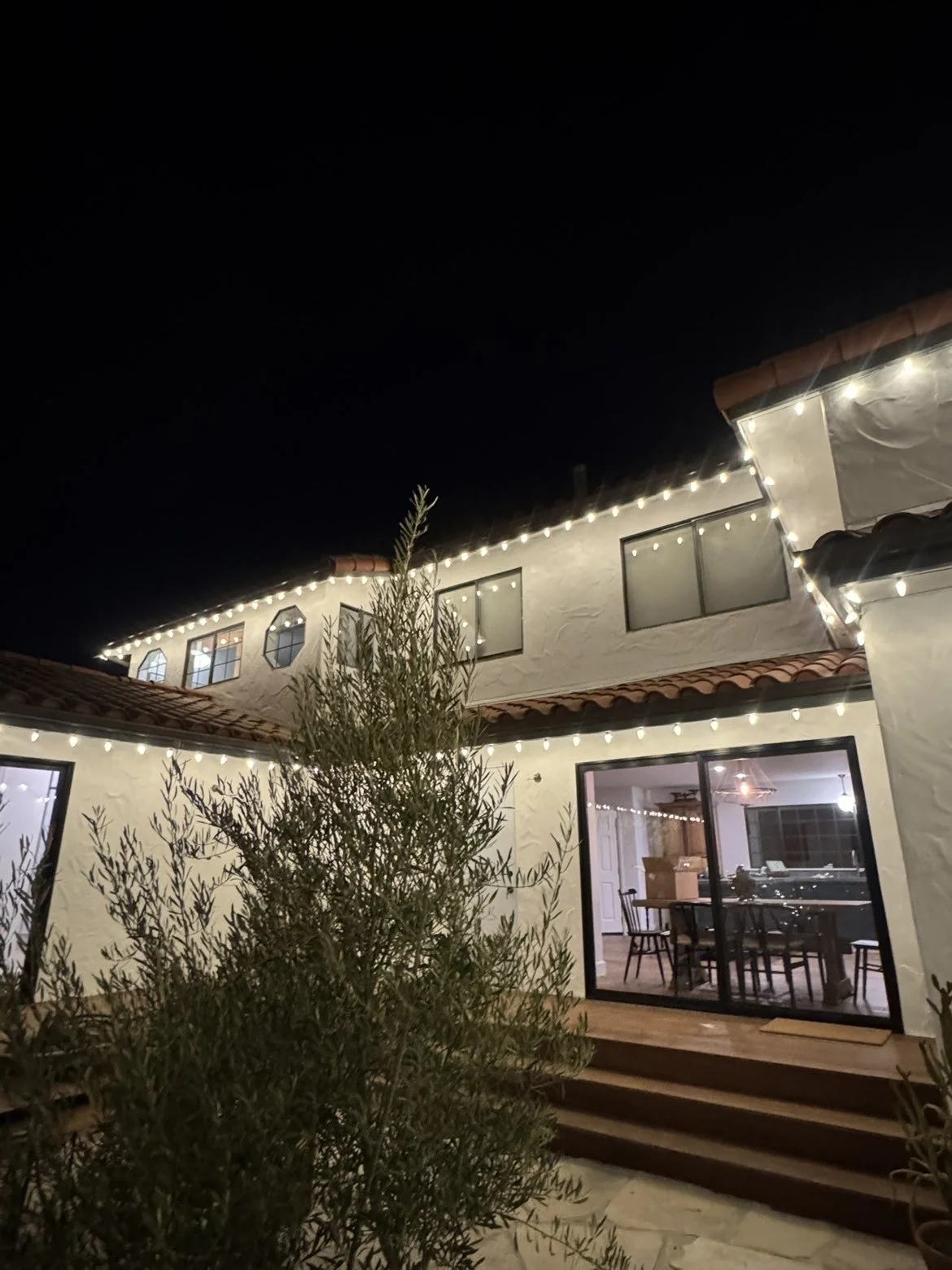 Nighttime view of a white house with string lights along the roofline, large glass sliding door revealing a dining area inside, and a small tree in the foreground.