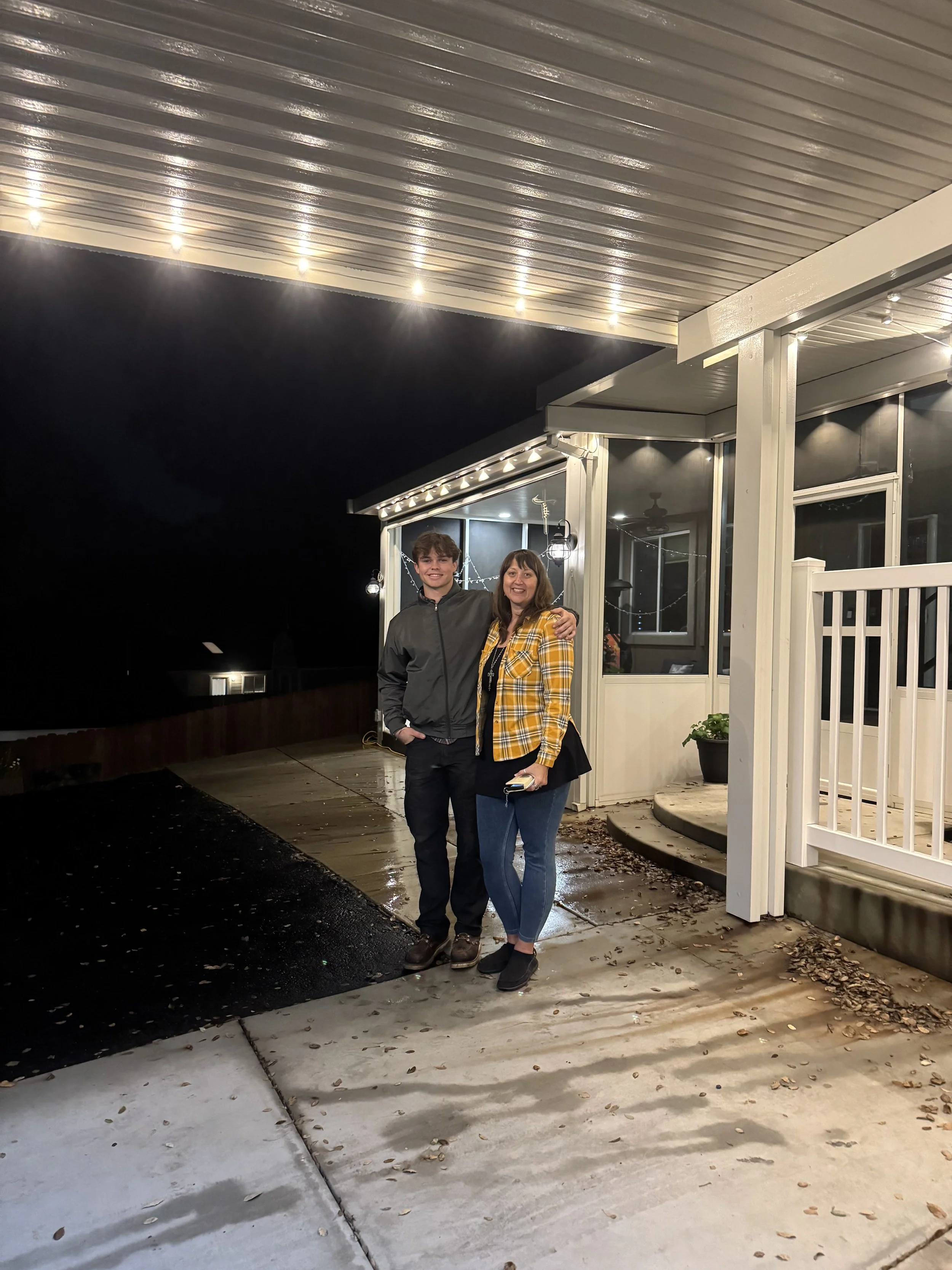 A woman and a young man standing together outside at night, smiling. They are on a porch or patio area with a metal roof and string lights.