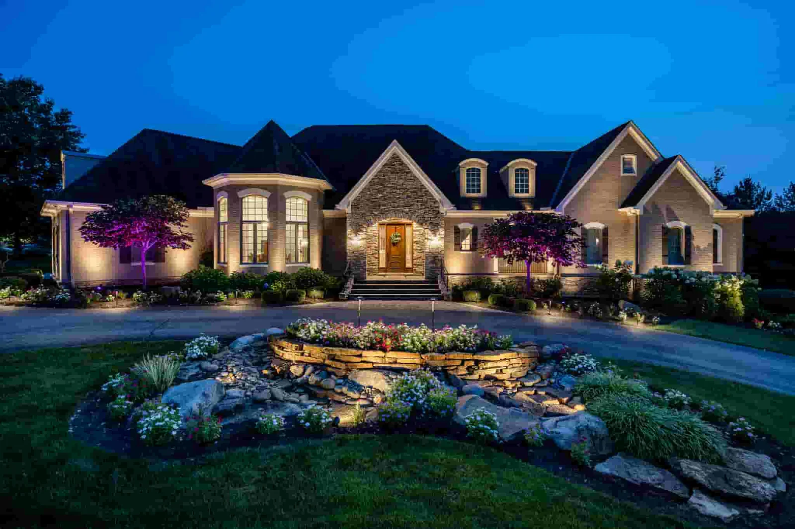 A well-lit house with a stone and brick facade at night, featuring large windows, a centered front door with a wreath, surrounded by landscaped garden beds, trees, and a curved driveway.
