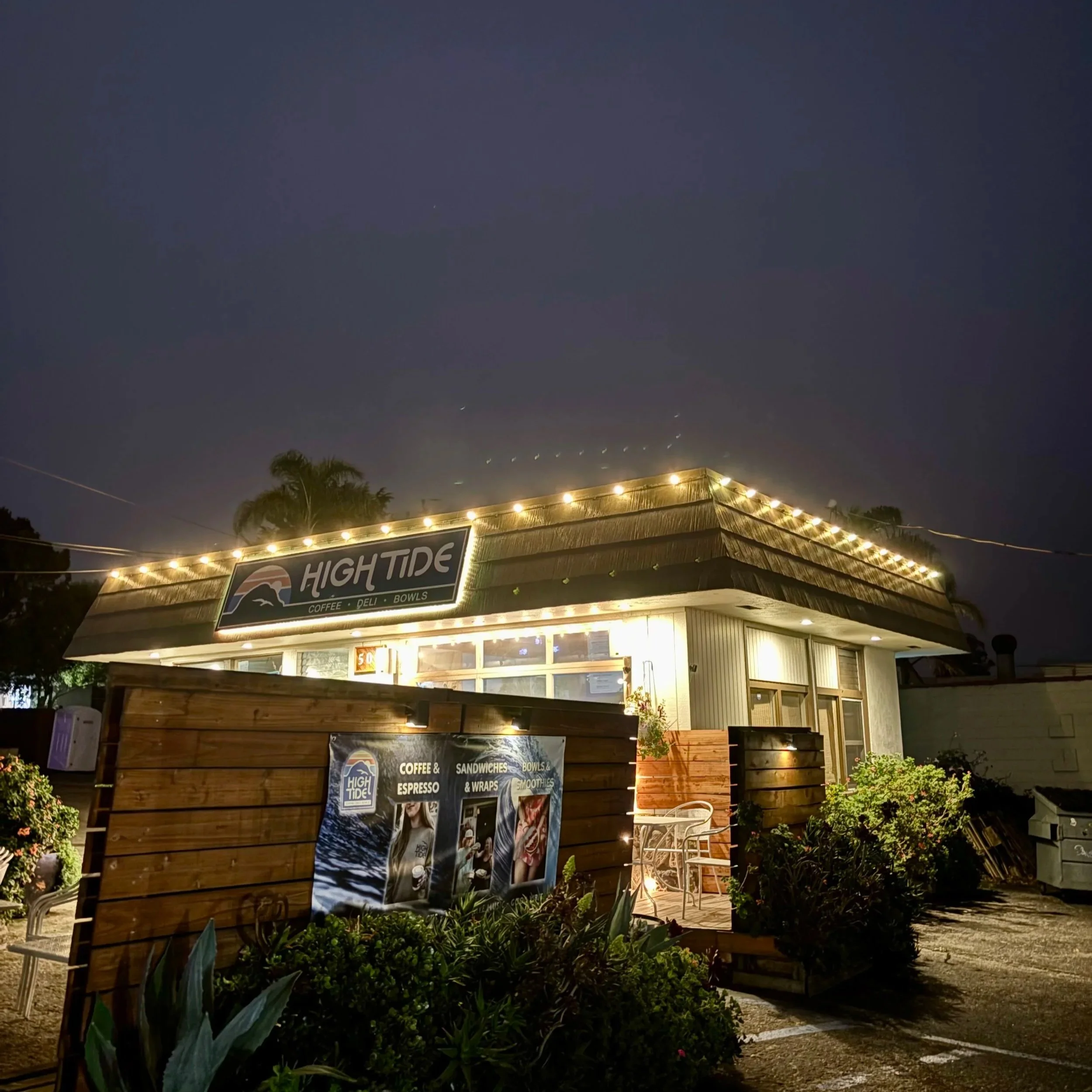 Night view of a small café named High Tide with string lights on the roof, outdoor seating, and promotional posters visible outside.