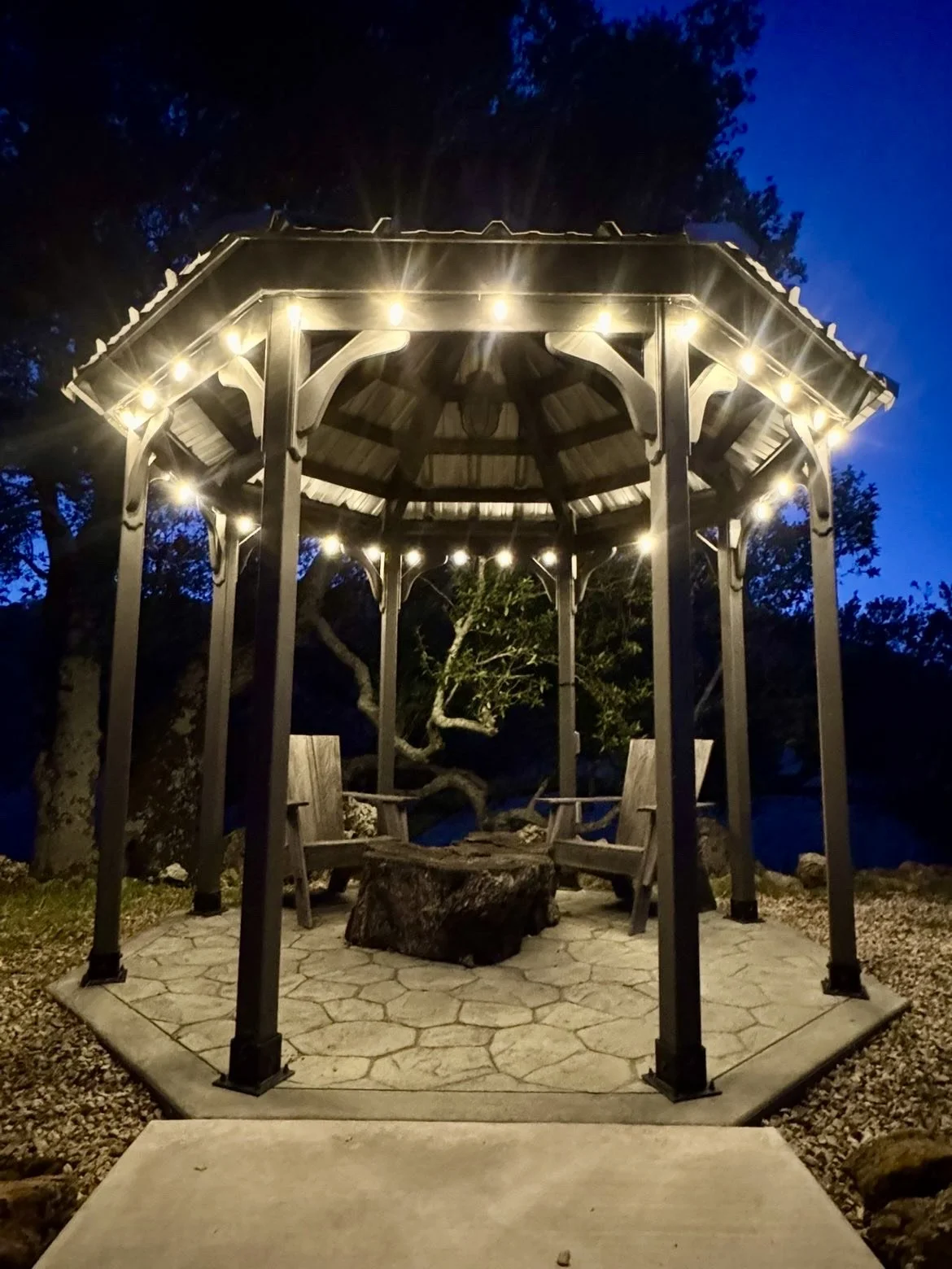 A lit gazebo with string lights, surrounded by trees at night, with two wooden chairs and a log table inside.