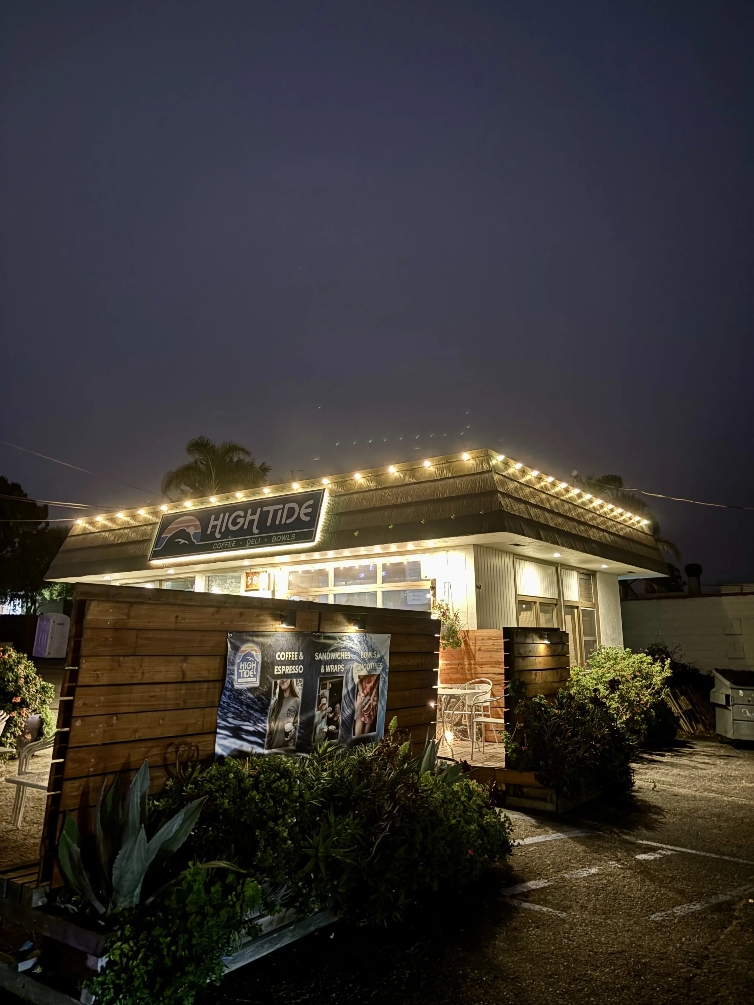 Night view of a small restaurant named 'High Tide' with a signboard advertising coffee, deli, bowls, sandwiches, wraps, and smoothies. The building is decorated with string lights, surrounded by plants and outdoor seating.
