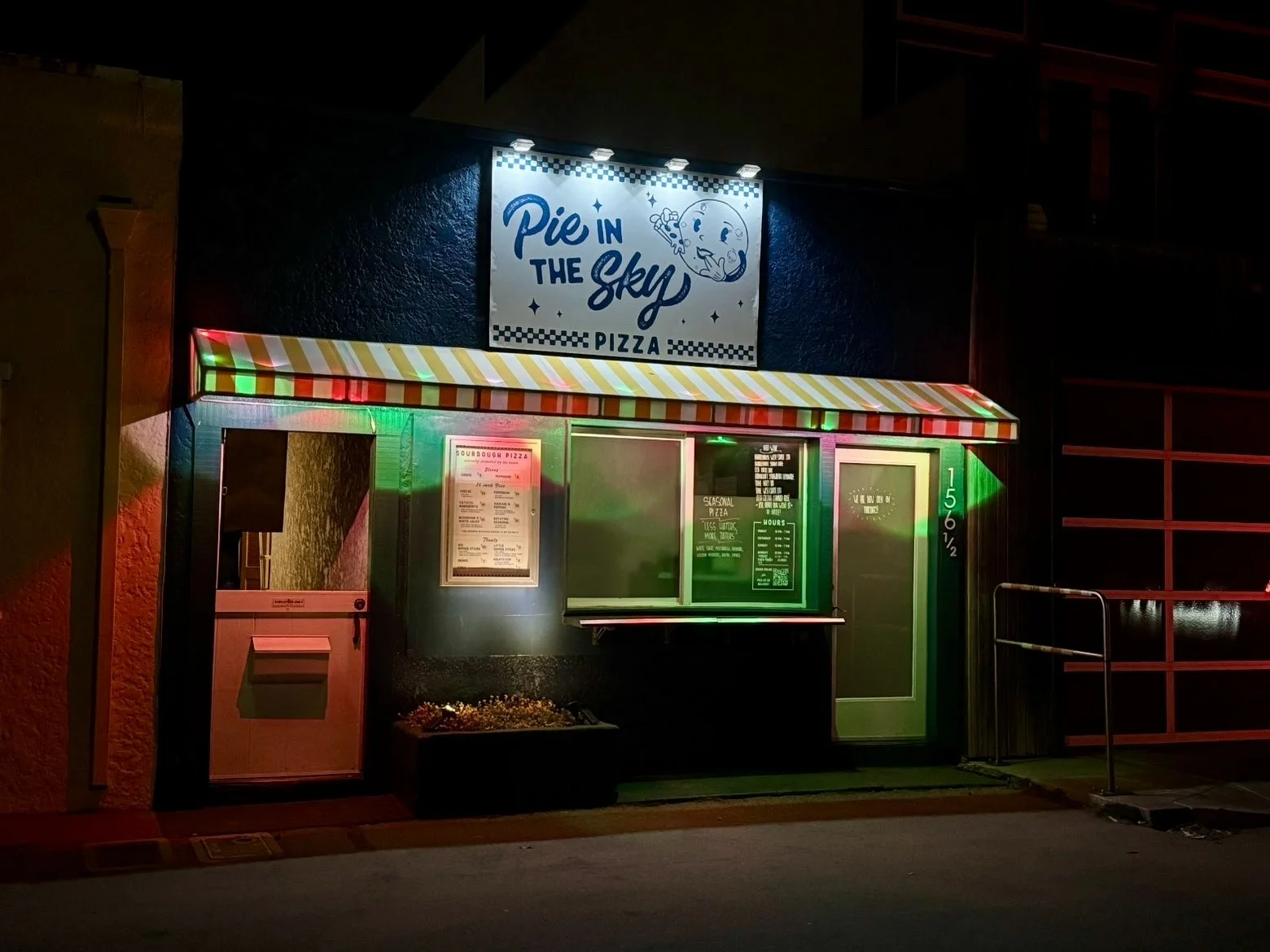 Nighttime view of a small pizza stand called "Pie in the Sky Pizza" with a colorful striped awning and illuminated menu signs.