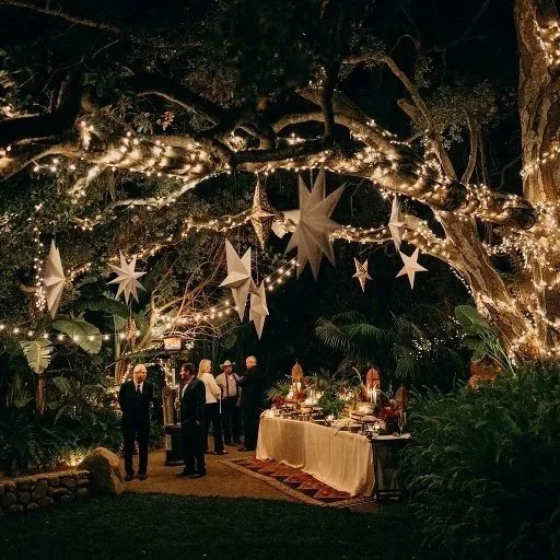 Nighttime outdoor party with hanging star and star-shaped paper lanterns, string lights, and a table of food and drinks, surrounded by trees and lush greenery.