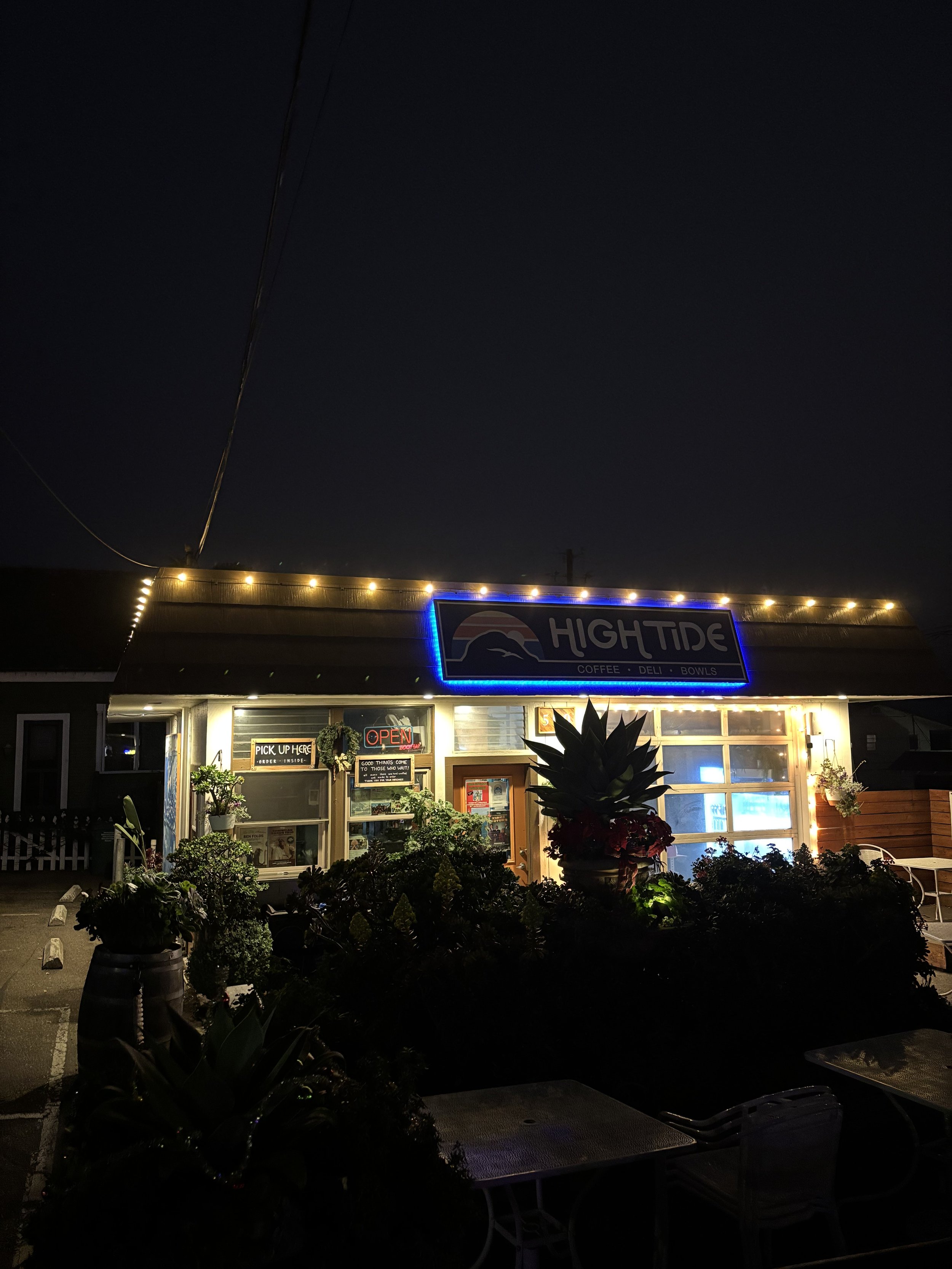 Nighttime photo of a small restaurant with a neon sign reading 'High Tide' above the entrance. The restaurant has string lights outlining the roof and large windows. There are signs indicating 'Open' and 'Pick Up Here,' along with various potted plan