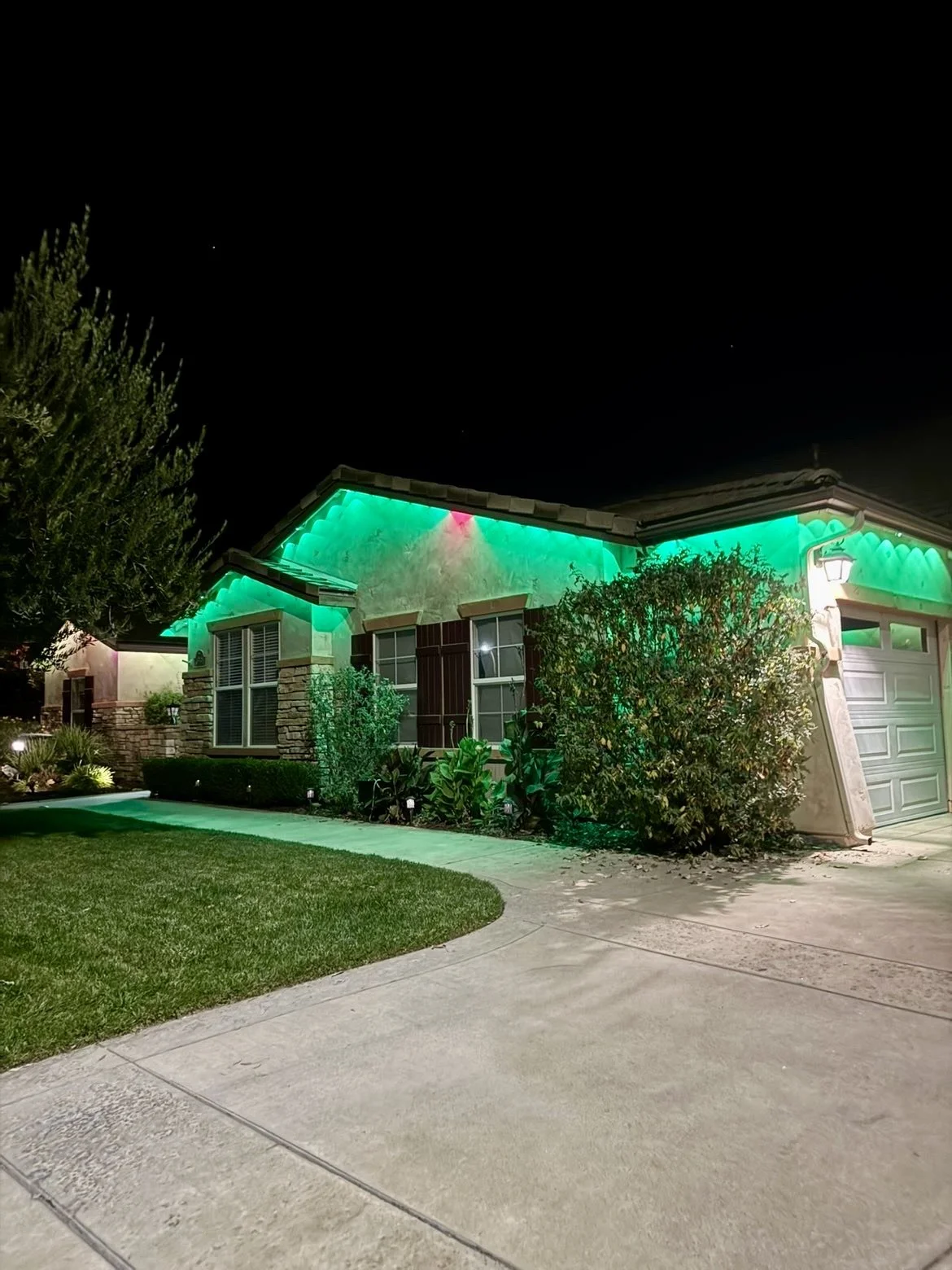 Night view of a house illuminated with green and pink outdoor Christmas or holiday lights, with a well-kept lawn and bushes in the front yard.