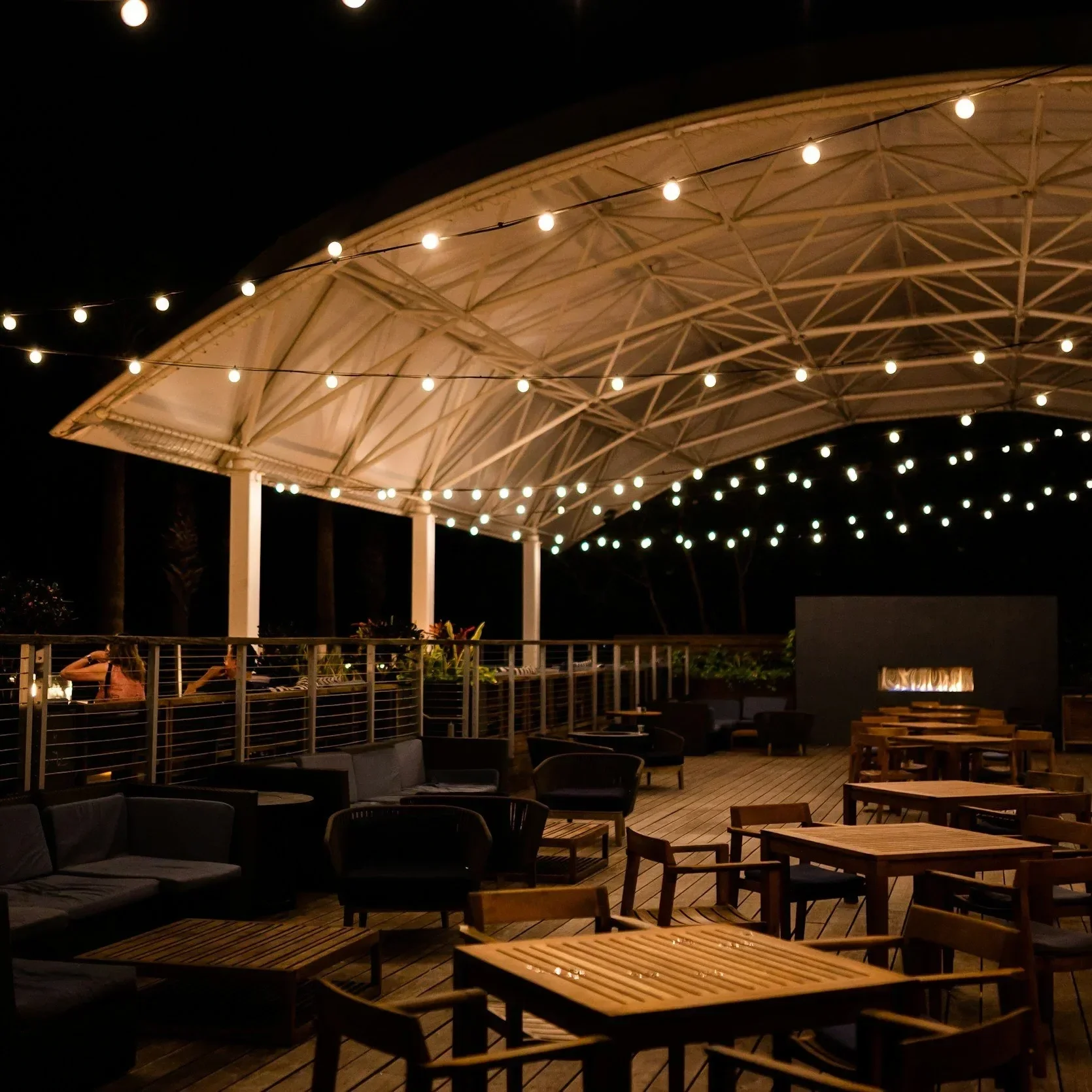 An outdoor rooftop seating area at night with string lights hanging from a canopy structure and wooden furniture.
