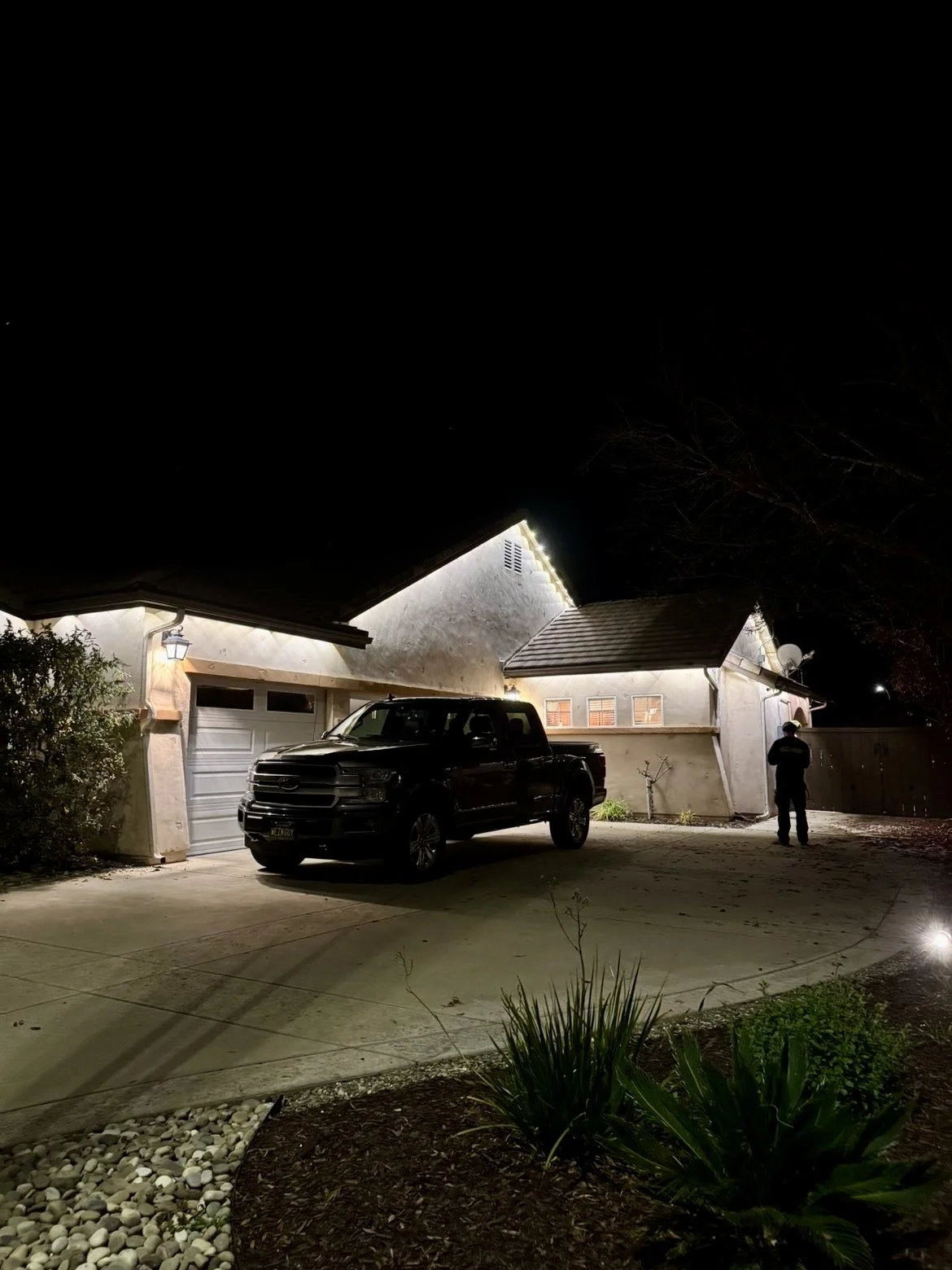 Nighttime view of a house with a garage, a black pickup truck parked in the driveway, and a person standing near the house. The house has exterior lighting along the roofline.