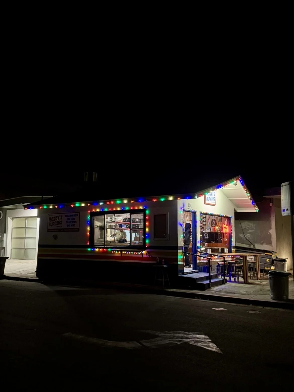 Night scene of a small food stand decorated with colorful string lights, with a person visible inside through the window, and an outdoor seating area with chairs and tables.