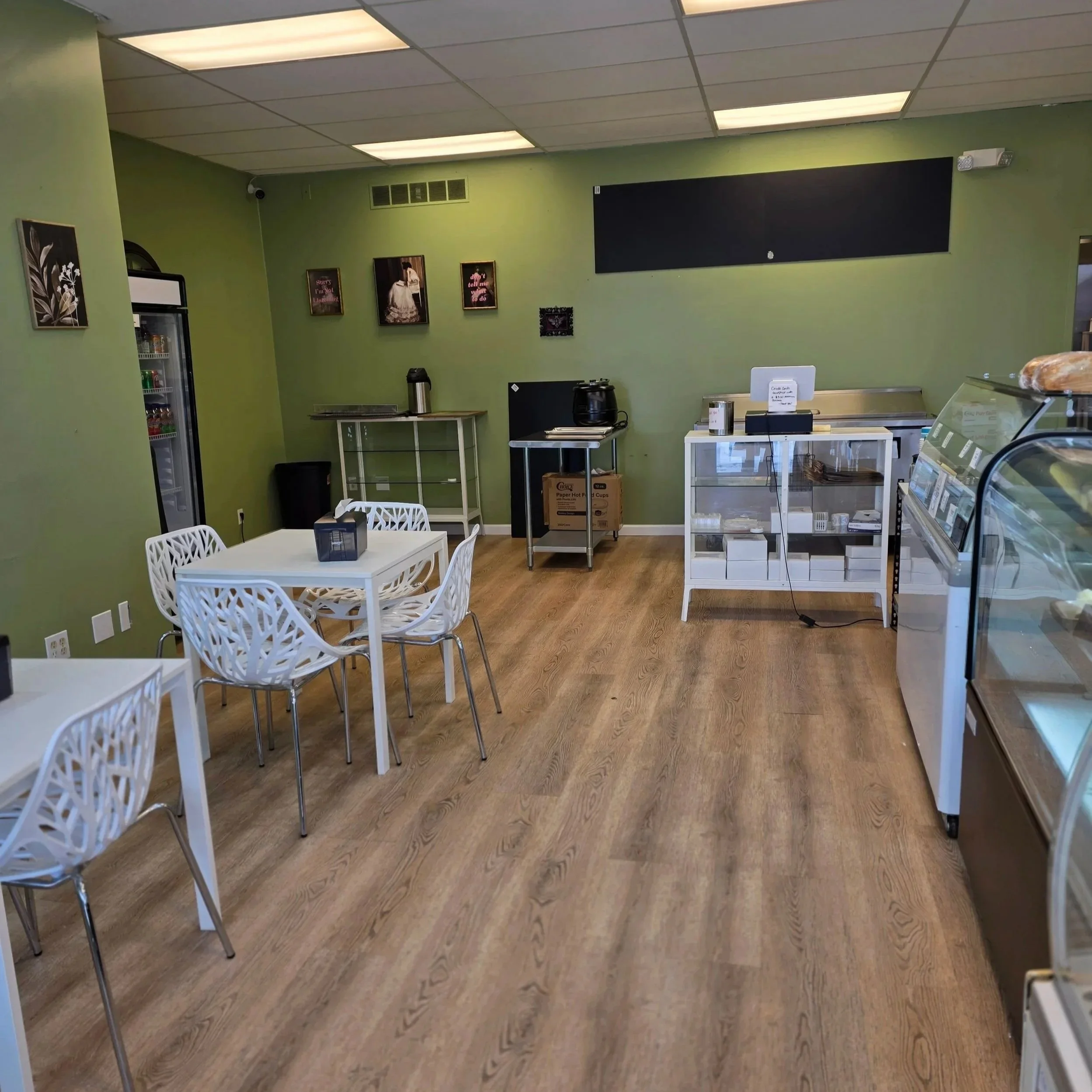 Interior of a small cafe with green walls, featuring white tables and chairs, a refrigerated display case, a counter with a cash register, and a few wall art pieces.