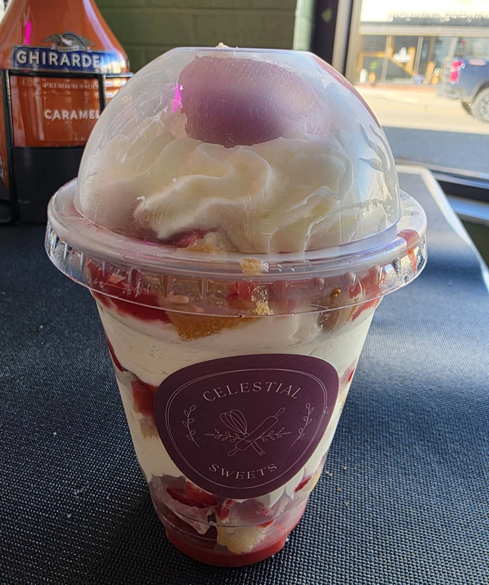 A clear plastic cup filled with strawberries, whipped cream, and a scoop of pink ice cream, topped with whipped cream inside a transparent lid, with a purple label reading 'Celestial Sweets.'