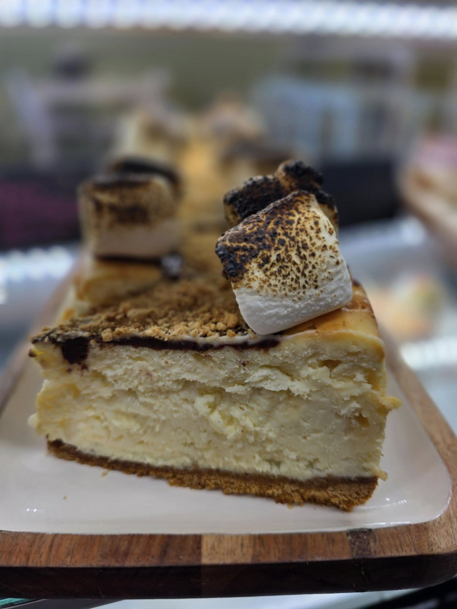 A slice of cheesecake topped with toasted marshmallows and graham cracker crumbs on a wooden serving board.