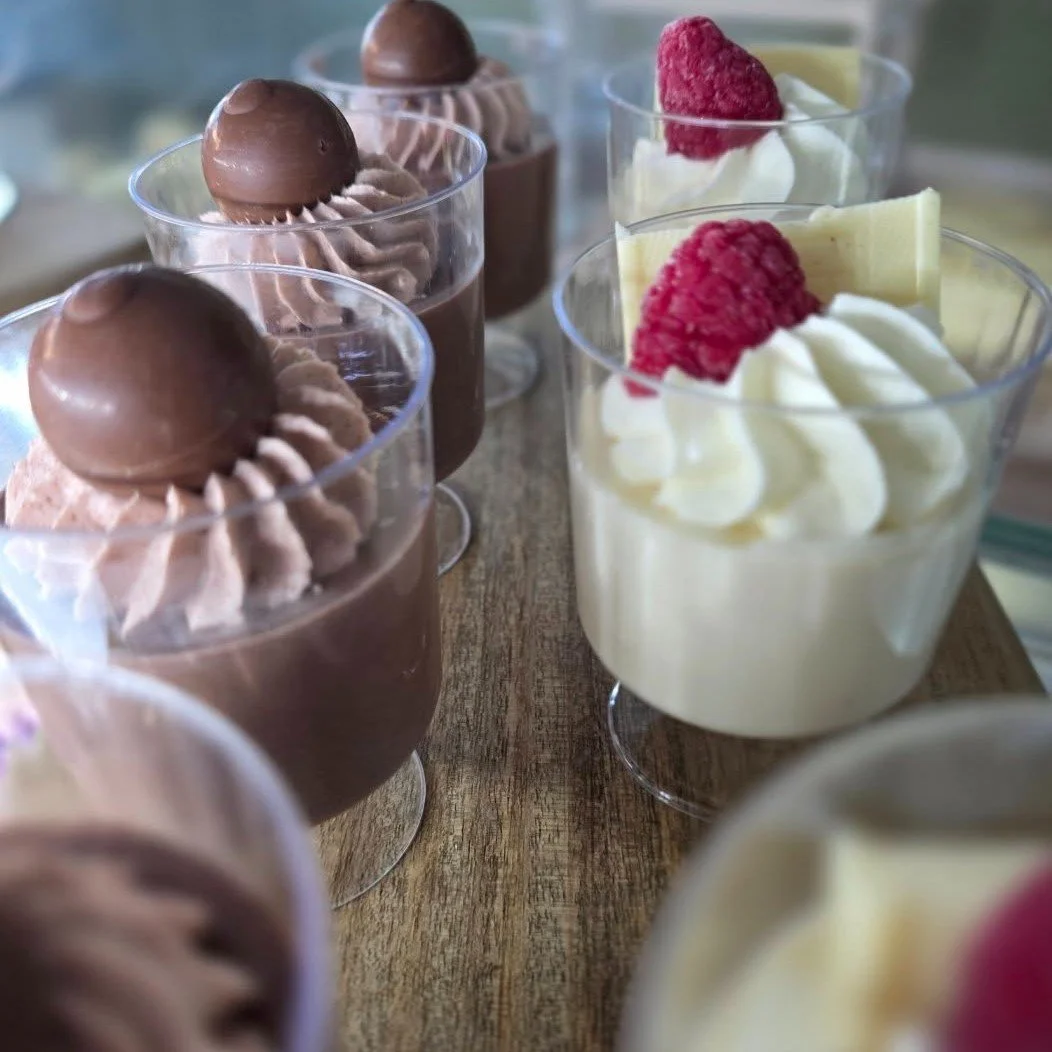 Clear glasses of chocolate mousse topped with chocolate spheres and whipped cream, and glasses of vanilla mousse topped with raspberries and white chocolate shavings, arranged on a wooden surface.