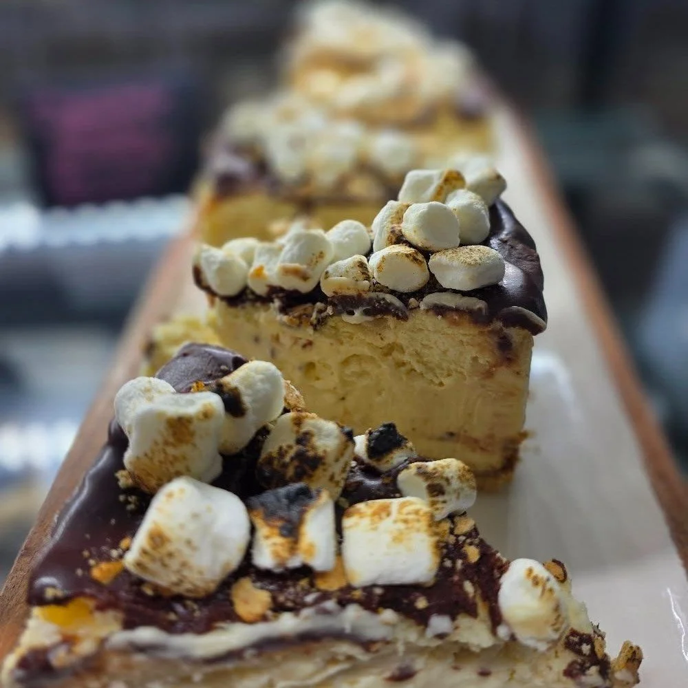 Close-up of a row of cheesecake slices topped with toasted marshmallows and chocolate drizzle on a white plate.
