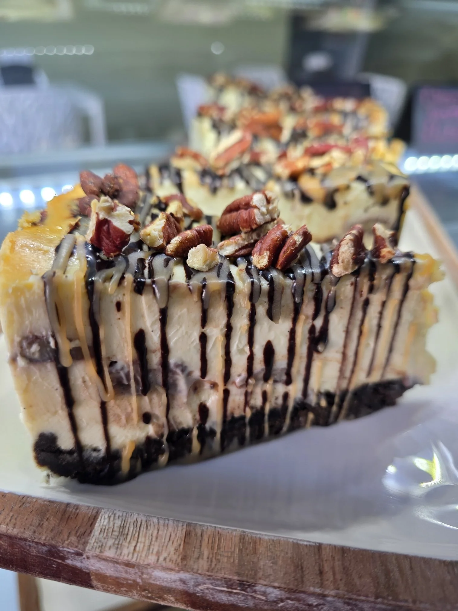 Close-up of a slice of cheesecake topped with chopped pecans and drizzled with chocolate and caramel sauces, with a similar slice in the background.