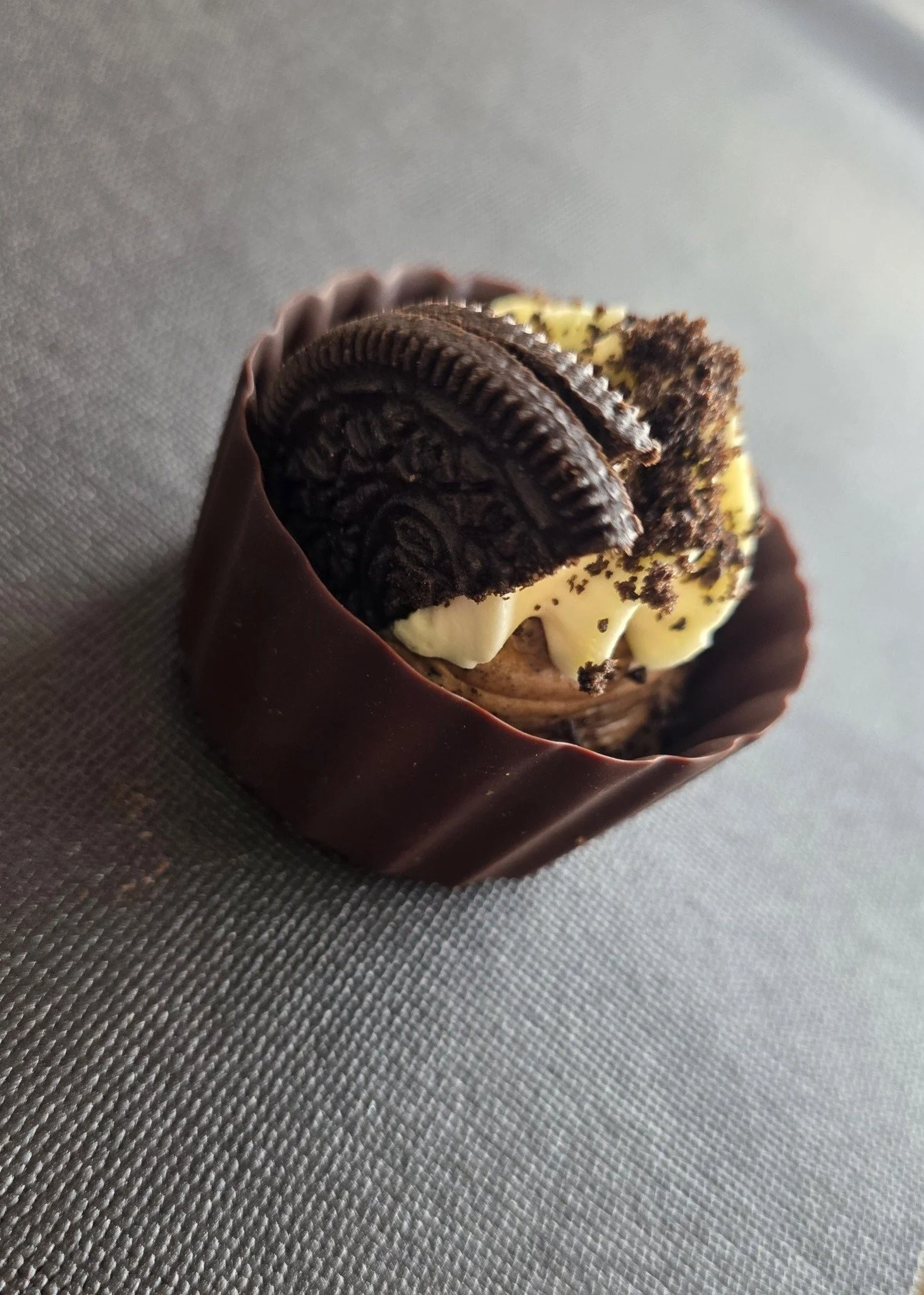 Close-up of a chocolate cupcake topped with white frosting, Oreo cookies, and crushed cookies in a dark chocolate wrapper on a textured gray surface.