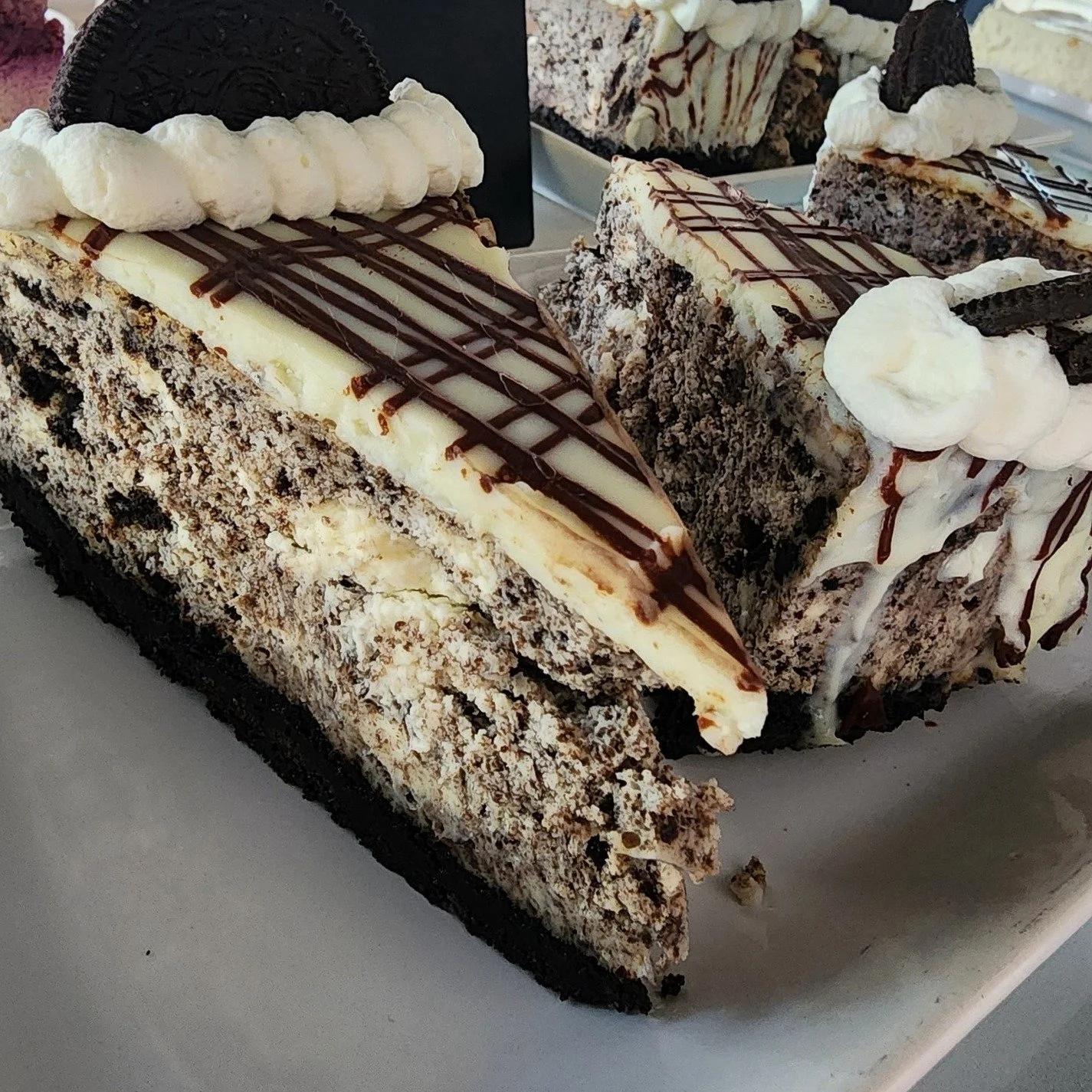 Slices of cookies and cream cheesecake with Oreo cookies, chocolate drizzle, and whipped cream decoration on a white plate.