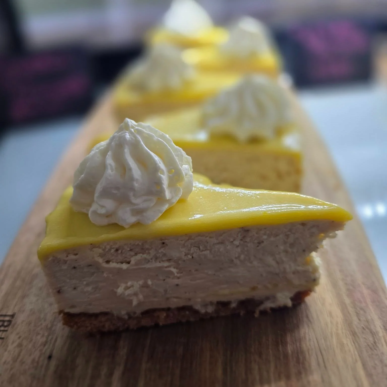 Close-up of a slice of lemon-flavored layered cake with whipped cream on top, placed on a wooden serving board, with more slices in the background.