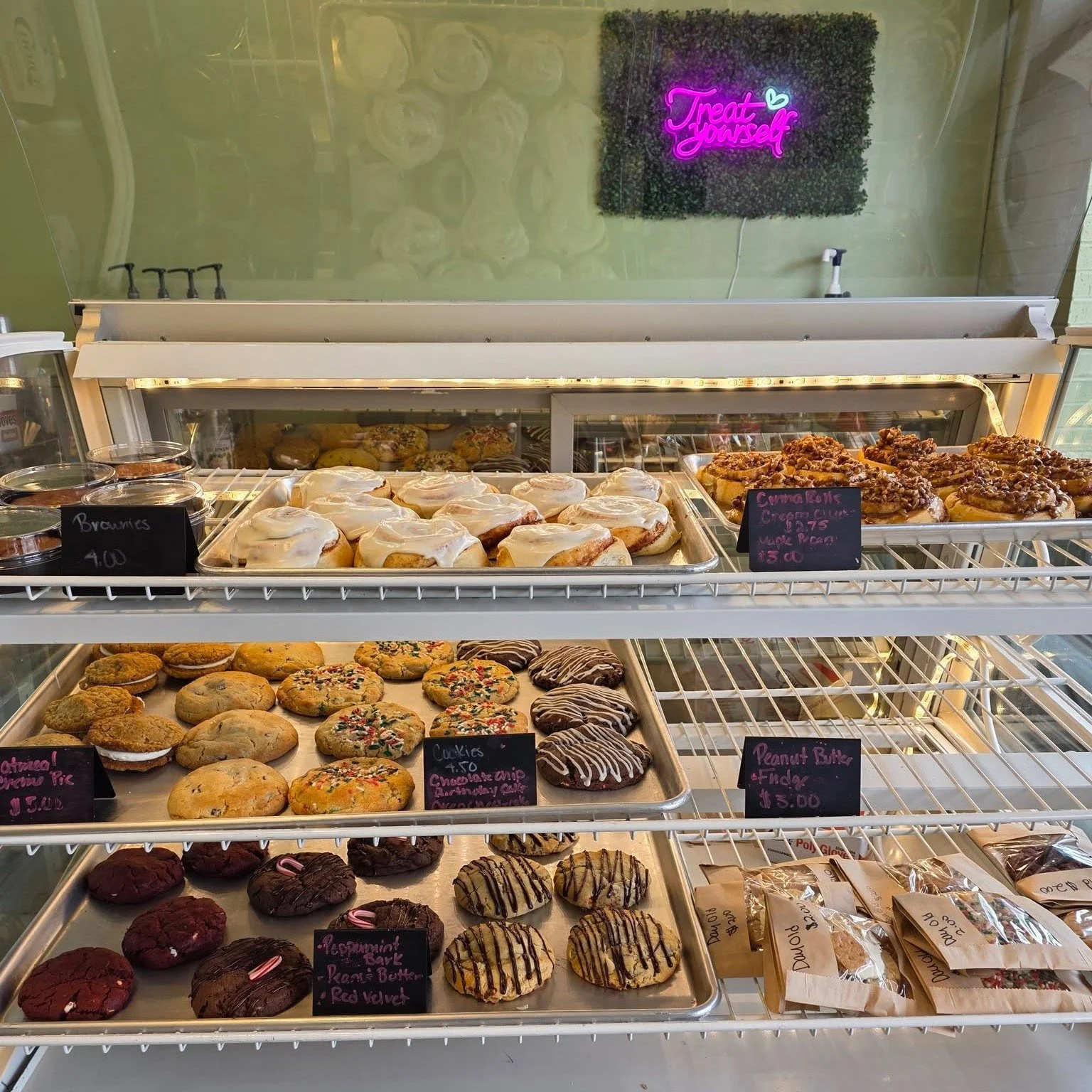 Display case with assorted baked goods including cookies, cinnamon rolls, brownies, and donuts, with a neon sign above that says "Treat Yourself".