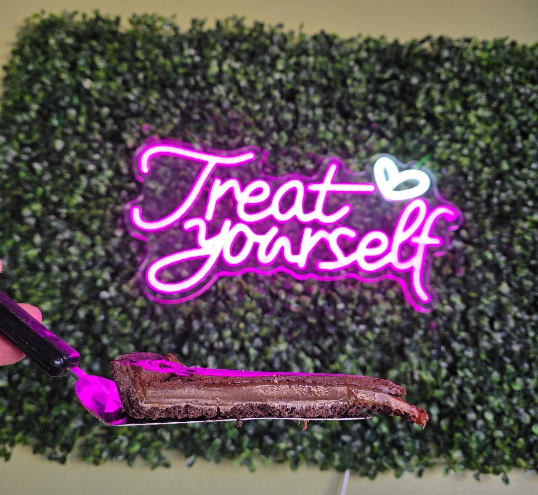 A chocolate layered dessert on a black-handled spatula in front of a neon sign that says 'Treat yourself' in pink and white, with a small white heart above the text, against a green plant wall background.
