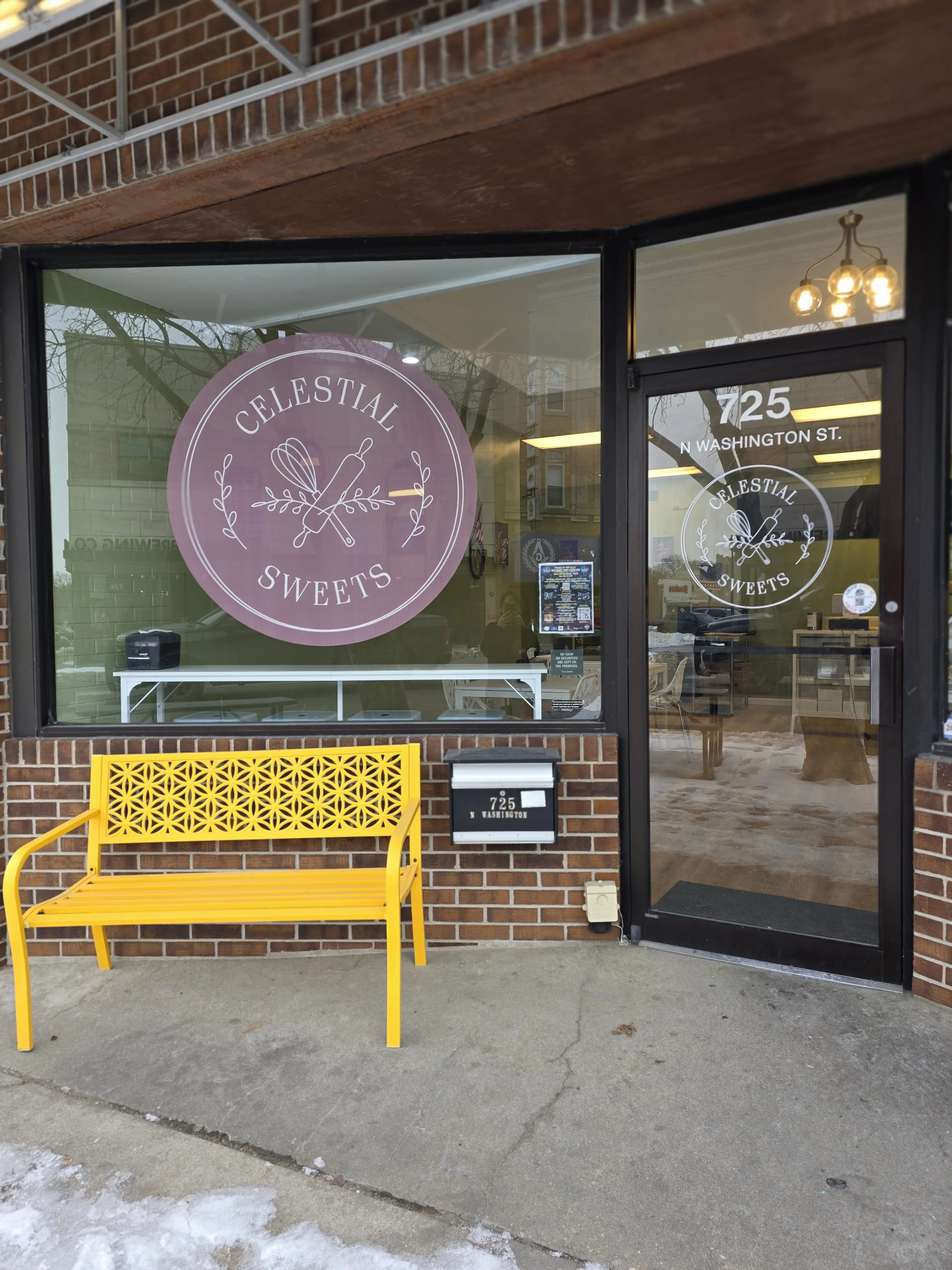 The storefront of Celestial Sweets with a large pink logo window featuring a whisk, rolling pin, and leaf design. A yellow bench is outside, and the street address 725 N Washington St. is displayed on the door and building.