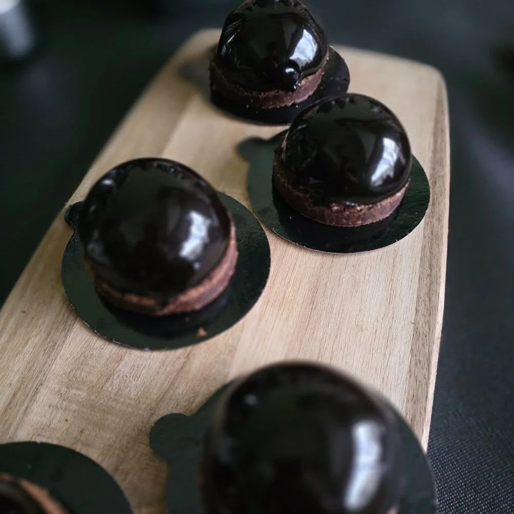 Four glossy dark chocolate dome desserts on black bases arranged on a wooden board.