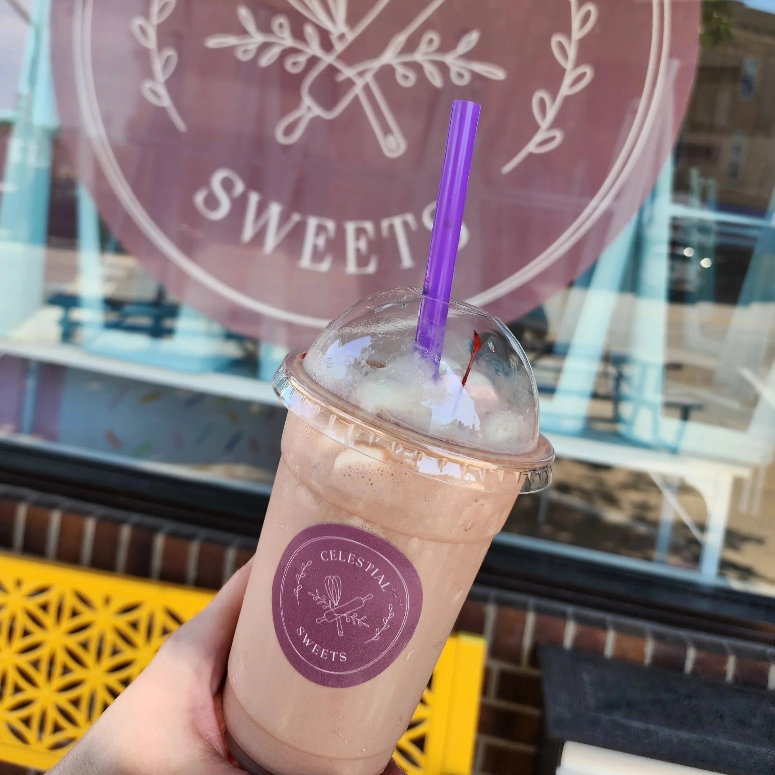 A hand holding a light pink smoothie in a clear plastic cup with a purple straw, in front of a window with a pink circular sign that reads "SWEETS". The sign has white decorative illustrations and text, with a yellow lattice bench and brick wall visible outside.
