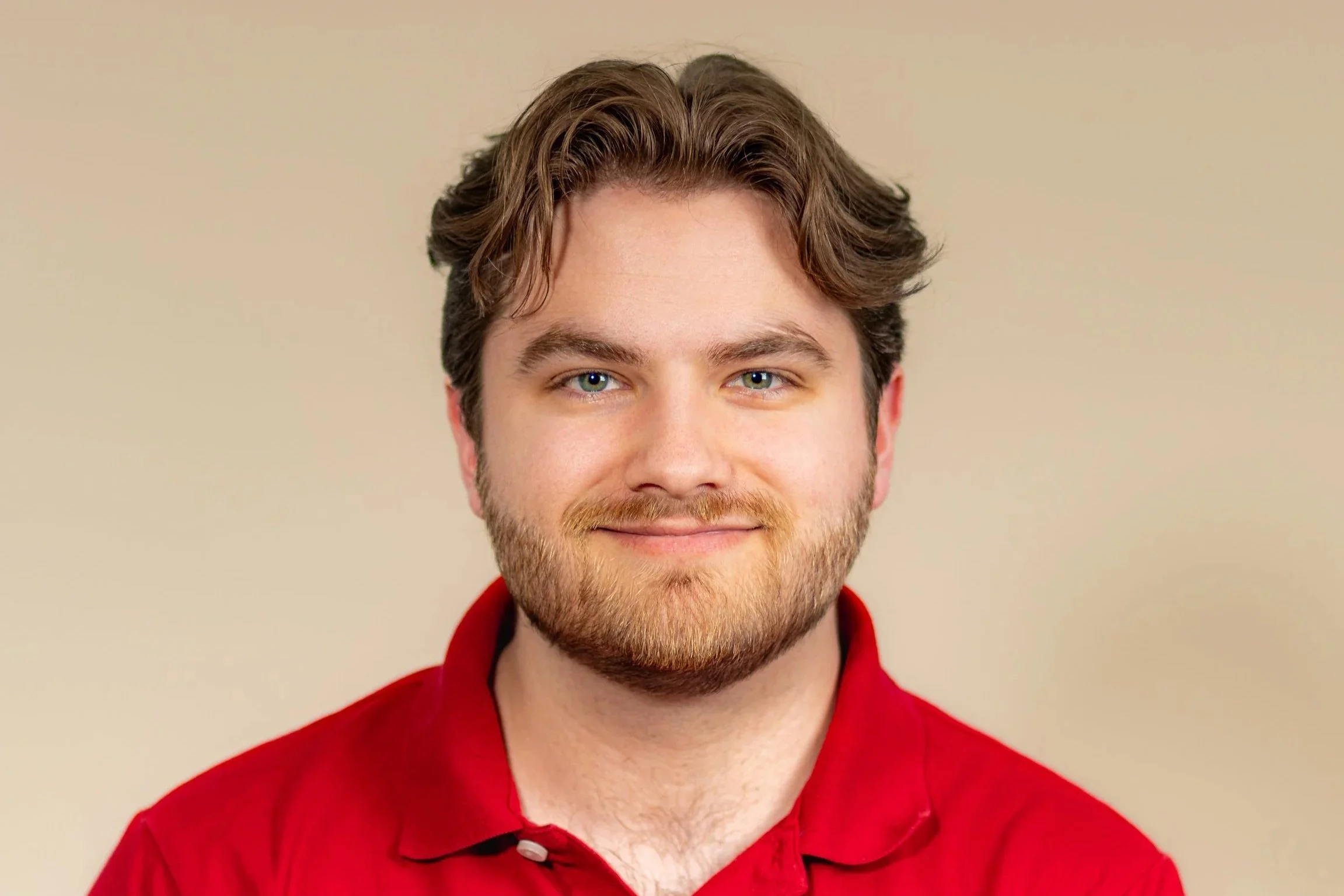 Portrait of a young man with light brown hair, blue eyes, and a beard, wearing a red collared shirt, smiling against a beige background.