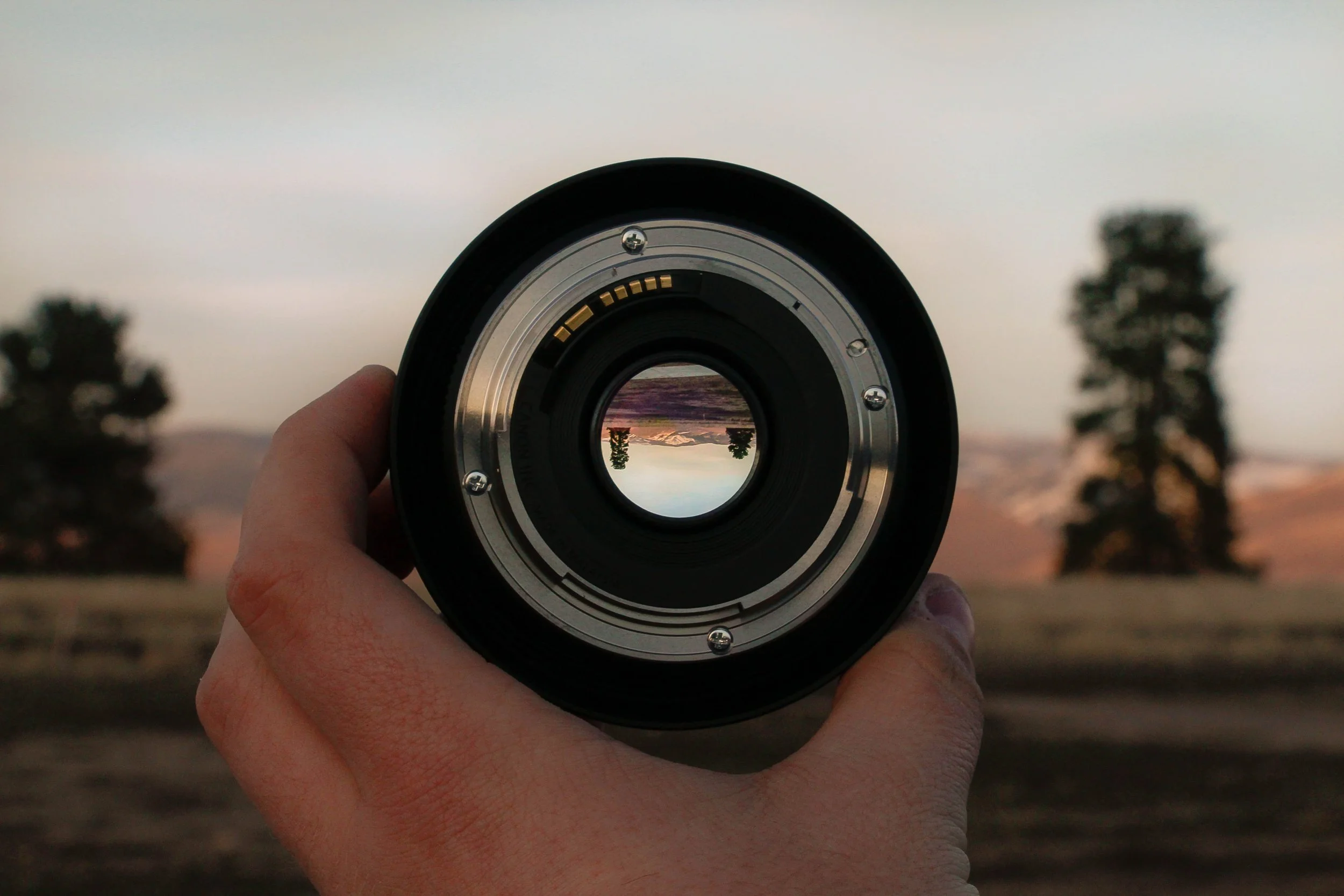 A person holding a camera lens in front of a scenic outdoor landscape at sunset, with trees and mountains visible through the lens.