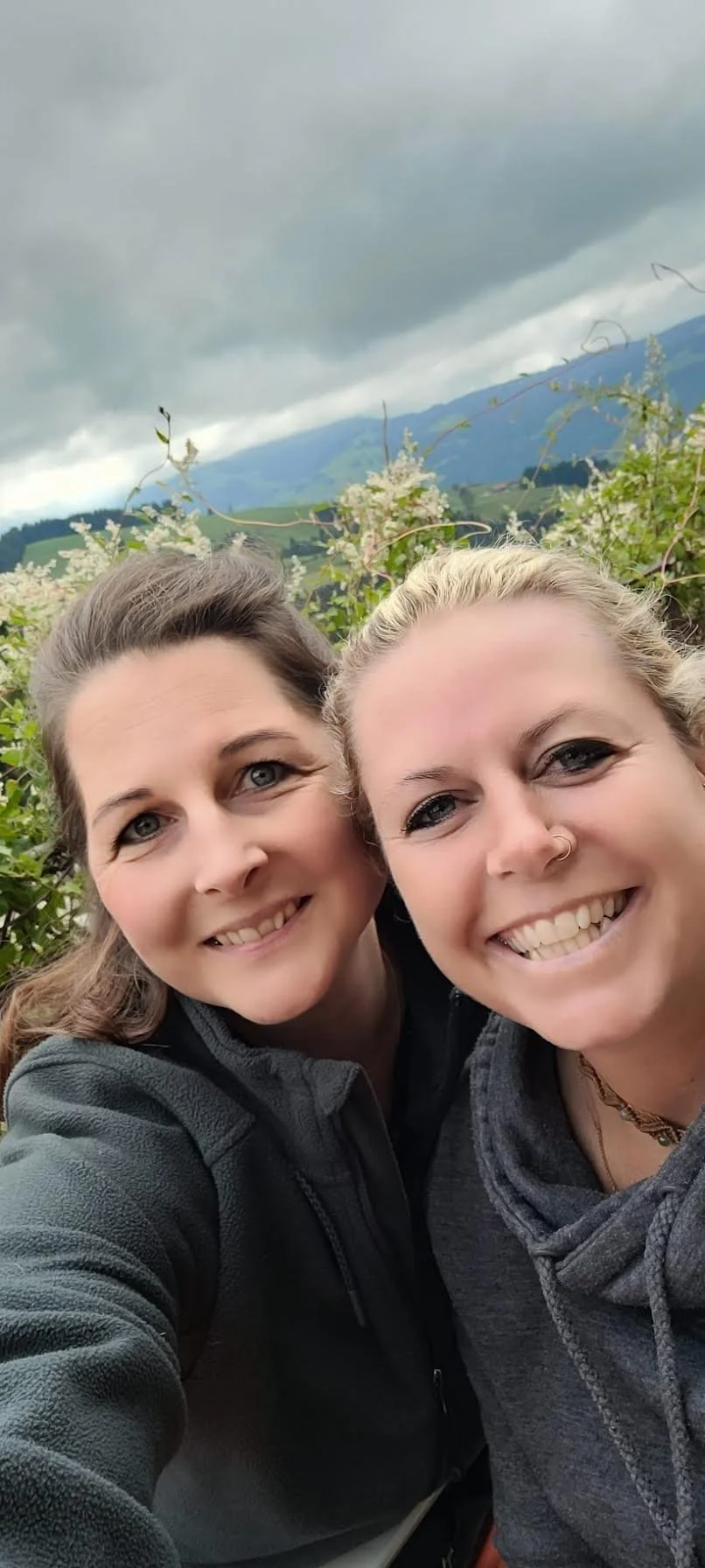 Zwei Frauen posieren im Freien mit Bergen, Wolken und Büschen im Hintergrund.