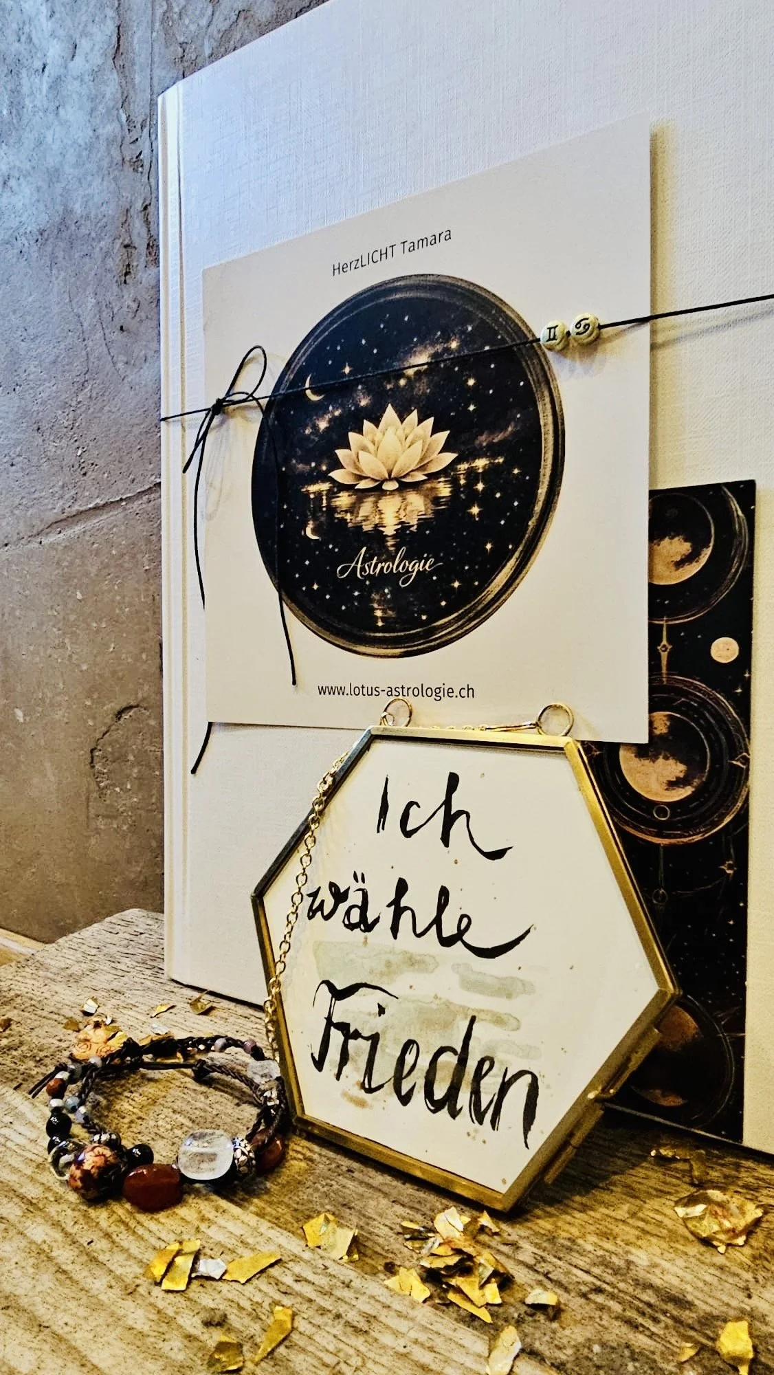 Ein Buch mit der Aufschrift 'Astrologie', ein weißer Uranus-Planeten-Schmuck, eine dekorative Schriftplatte mit der Aufschrift 'Ich wähle Frieden', ein Armband aus bunten Perlen und Goldfolie auf einem Holztisch.