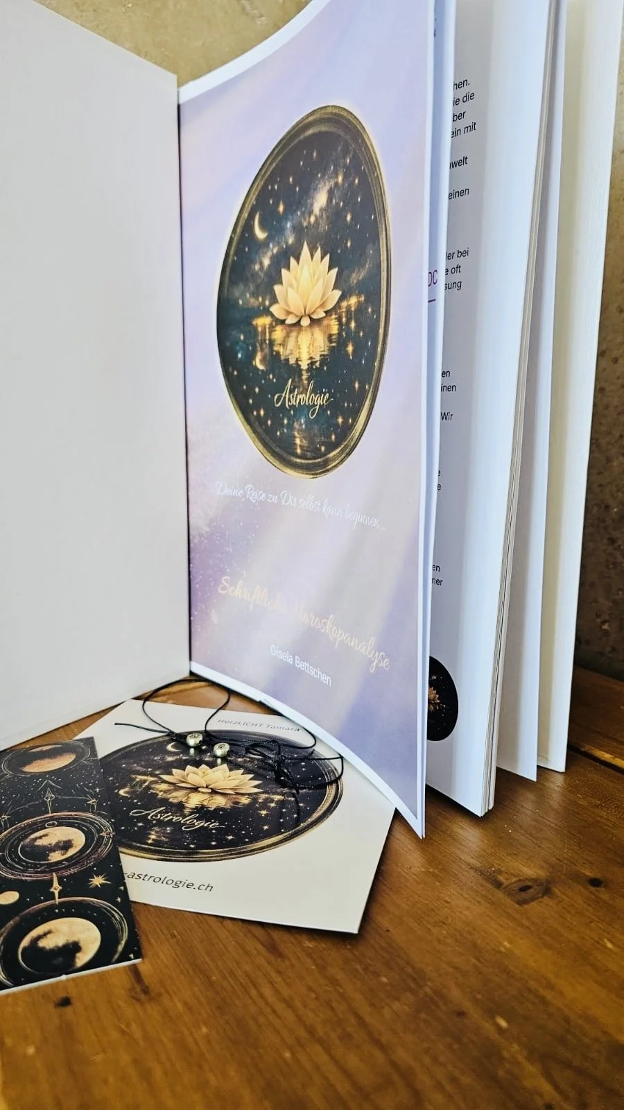 Ein aufgeschlagenes Buch über Astrologie mit einem Lotusblumen-Design auf dem Cover, mehreren Visitenkarten und einem Talisman mit Sternen- und Mondenmotiven auf einem Holztisch.