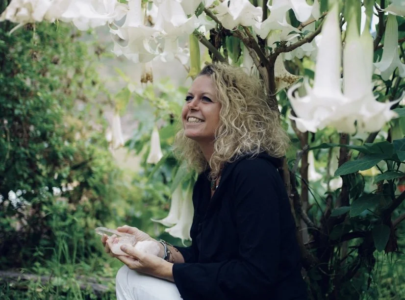 Frau mit lockigem blondem Haar sitzt im Garten neben weißen Blumen.