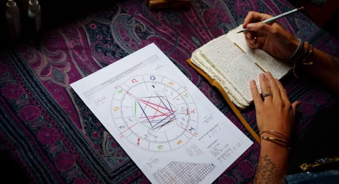 Person schreibt in ein Notizbuch, neben ihr astrologisches Diagramm auf einem Blatt Papier auf einem bunten Teppich.
