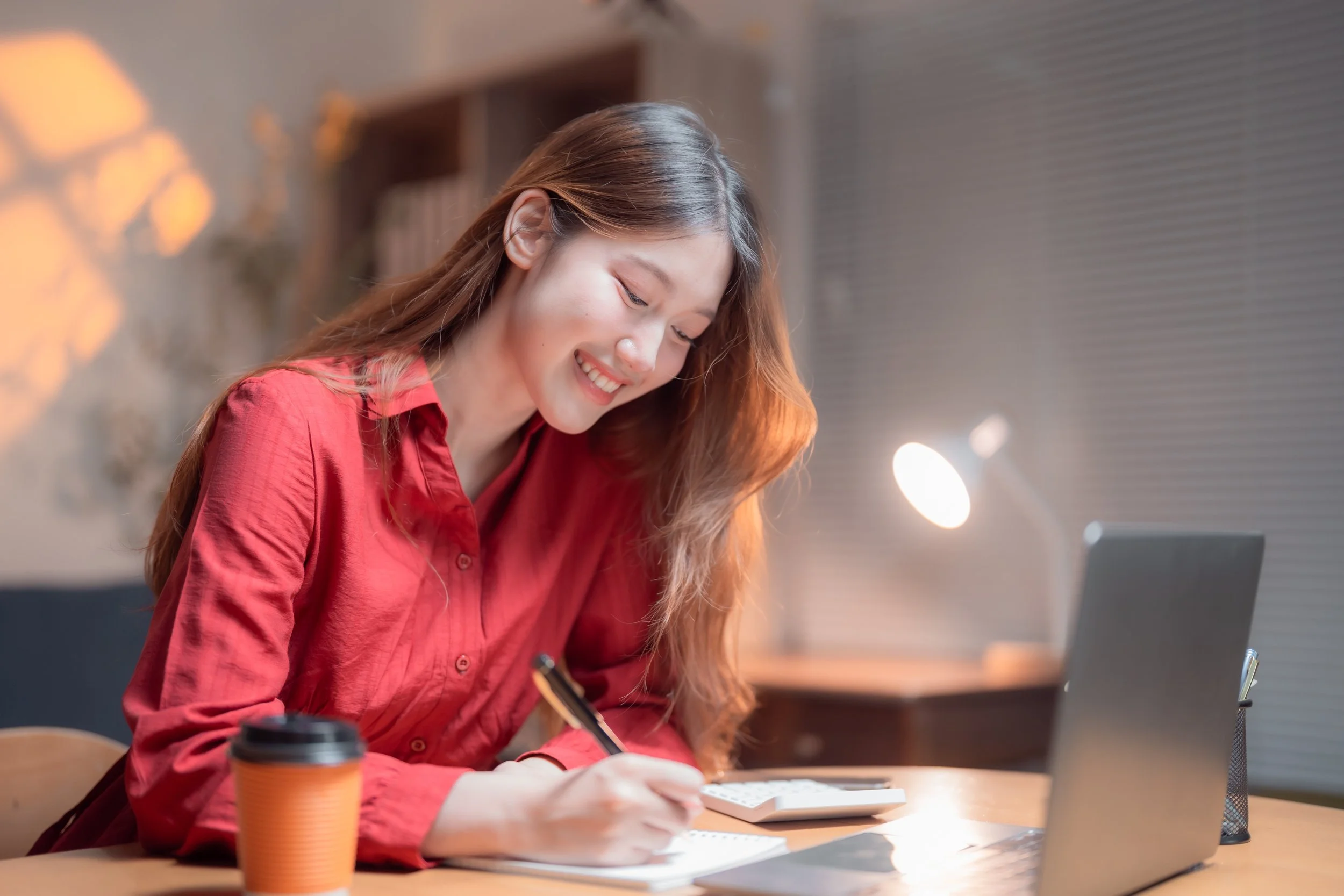 A young woman with long reddish-brown hair wearing a red shirt, sitting at a desk in a modern office or cafe, smiling while writing in a notebook with a black pen. There is a disposable coffee cup and a laptop on the desk. The background shows a shelf with books and a window with blinds.