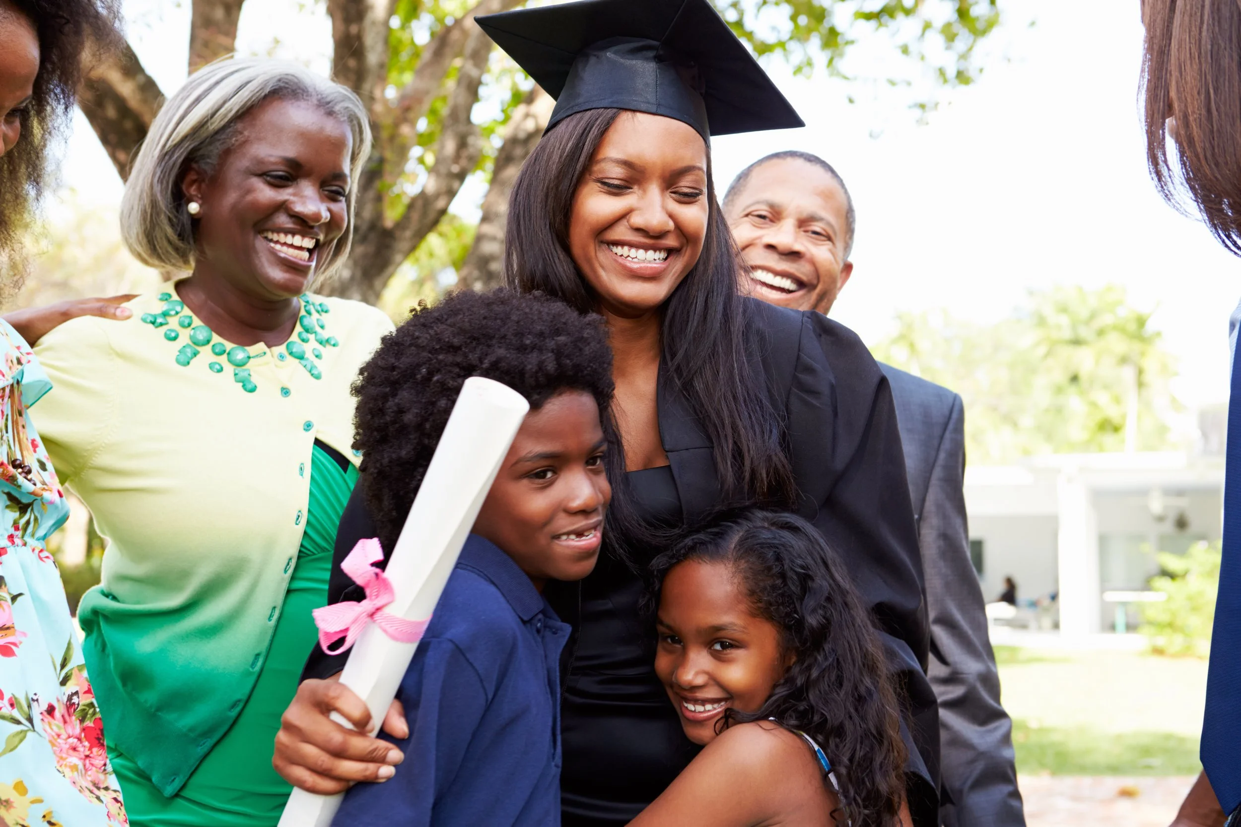 Group of smiling people, including children and adults, outdoors during a graduation celebration, with a woman in a cap and gown.