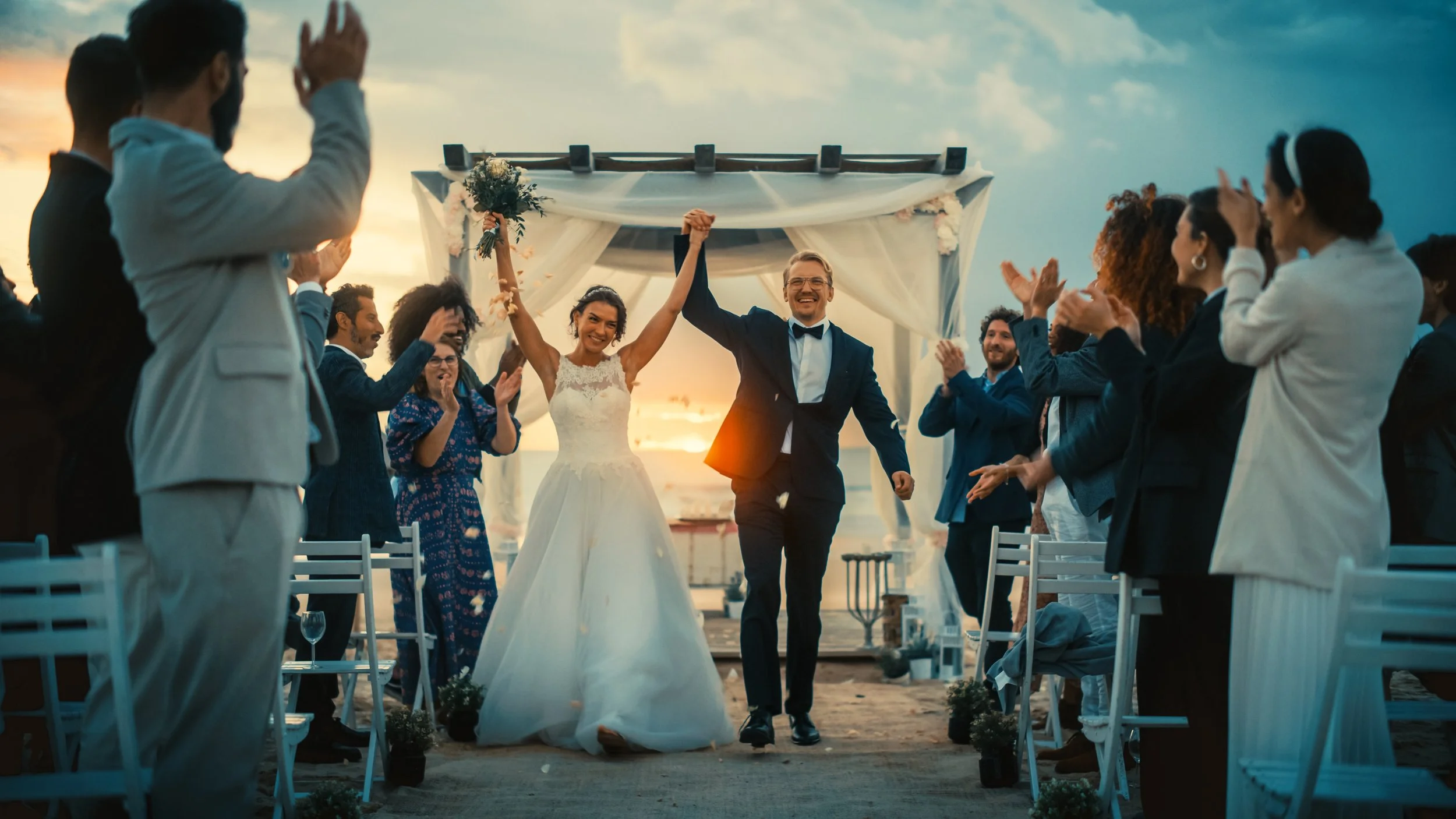 A newly married couple is walking down the aisle at sunset, holding hands and smiling, surrounded by friends and family clapping and celebrating.