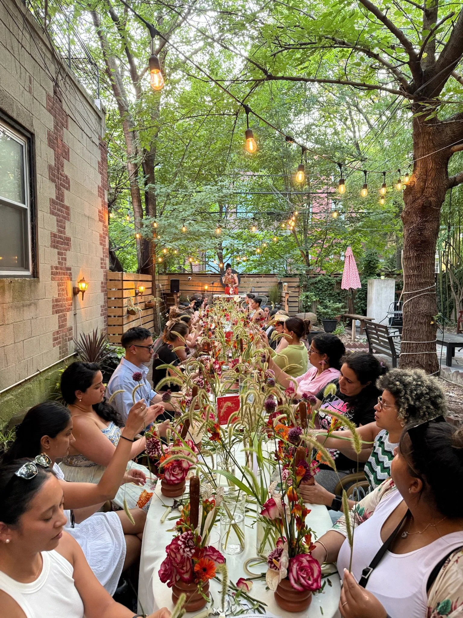 A long outdoor table with people seated on both sides, decorated with colorful flower arrangements in small pots. Overhead string lights are hanging, and a woman is speaking or performing in front of the group. The setting is in a garden area surrounded by trees and a brick wall.