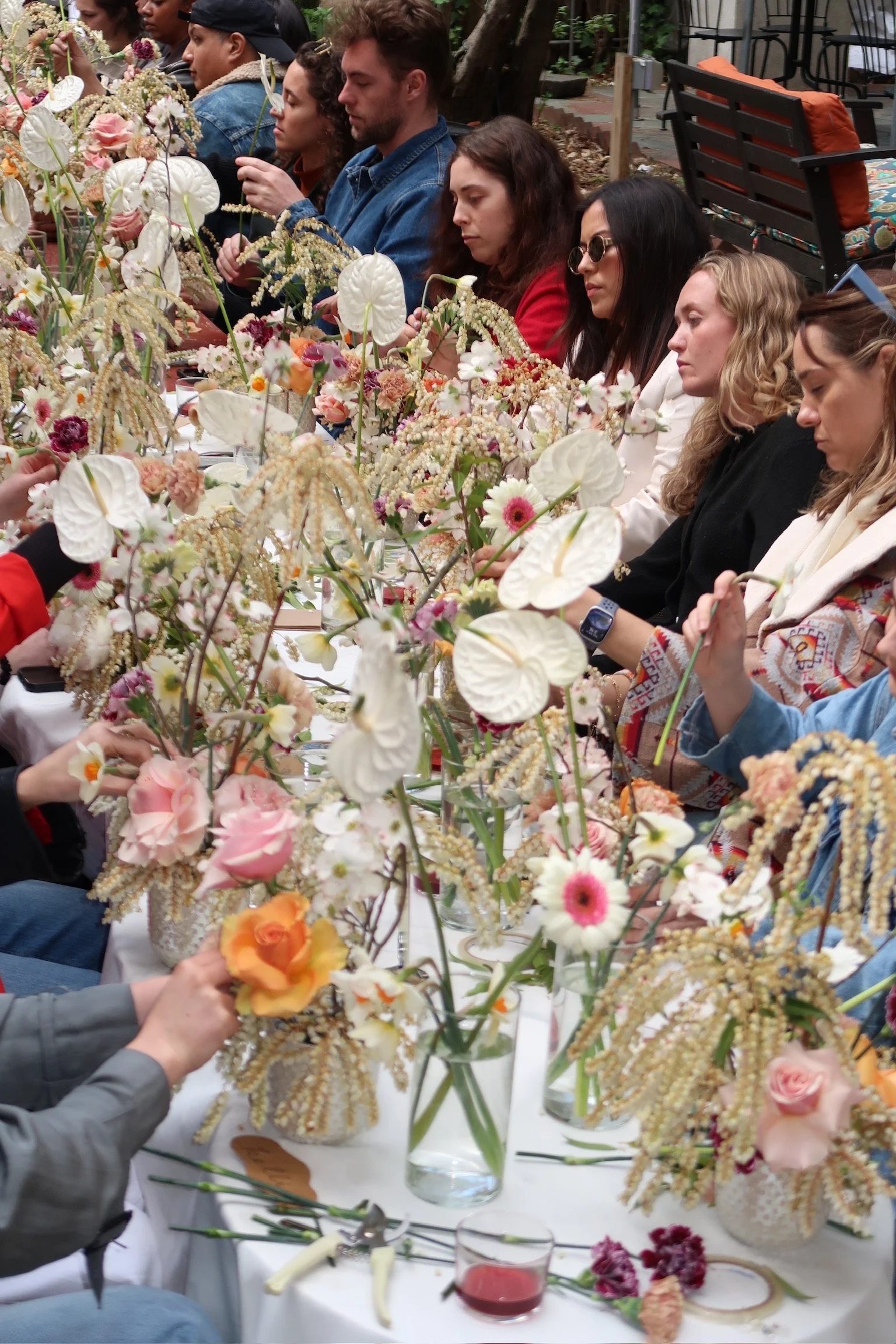People sitting at a long table with floral arrangements, engaged in a flower arranging activity outdoors.