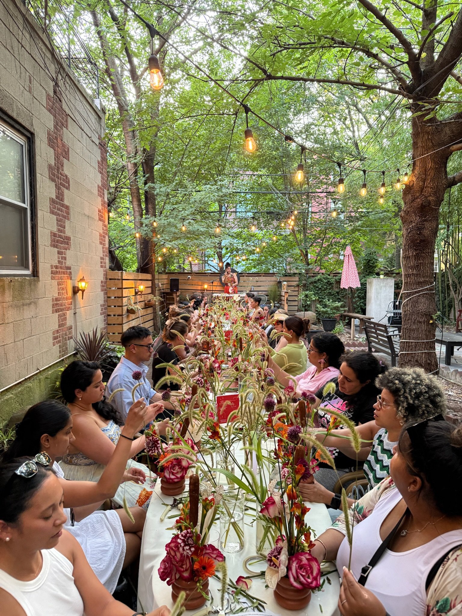 People sitting at a long outdoor table with floral arrangements, surrounded by trees and string lights