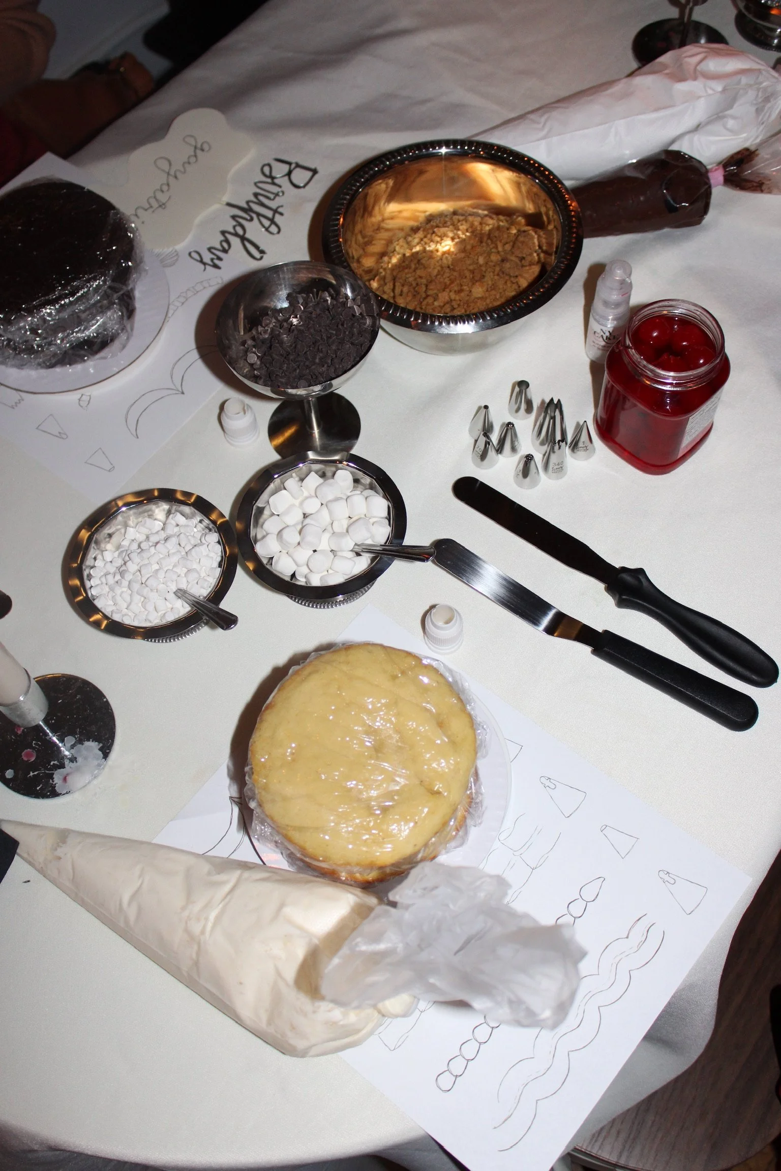 Bake decorating tools and ingredients on a table, including piping tips, marshmallows, chocolate chips, candy cherries, a cake with yellow icing, and baking drawings.