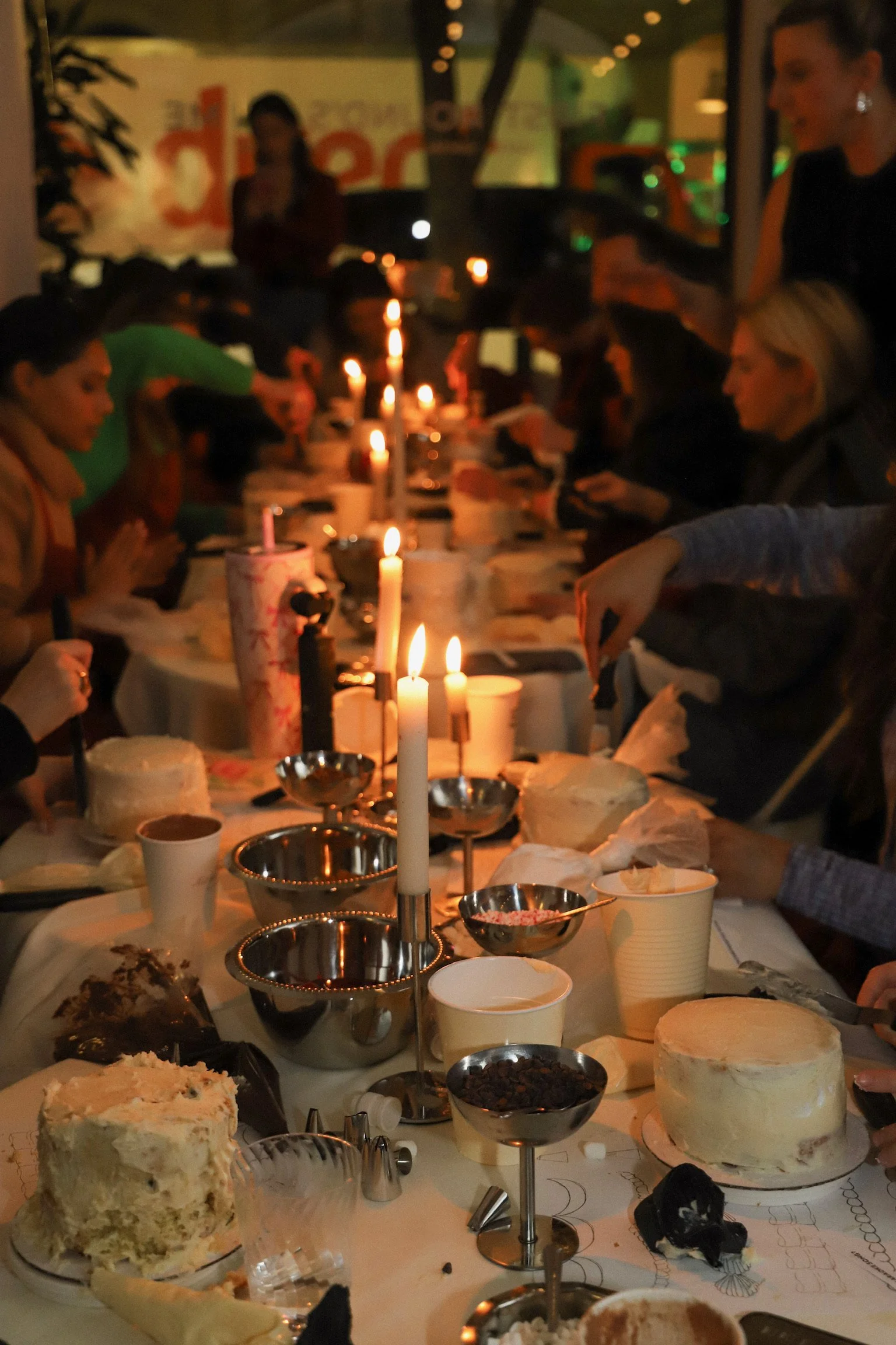 A large table with birthday cakes, candles, and desserts, surrounded by people celebrating at night in a dimly lit restaurant or party setting.