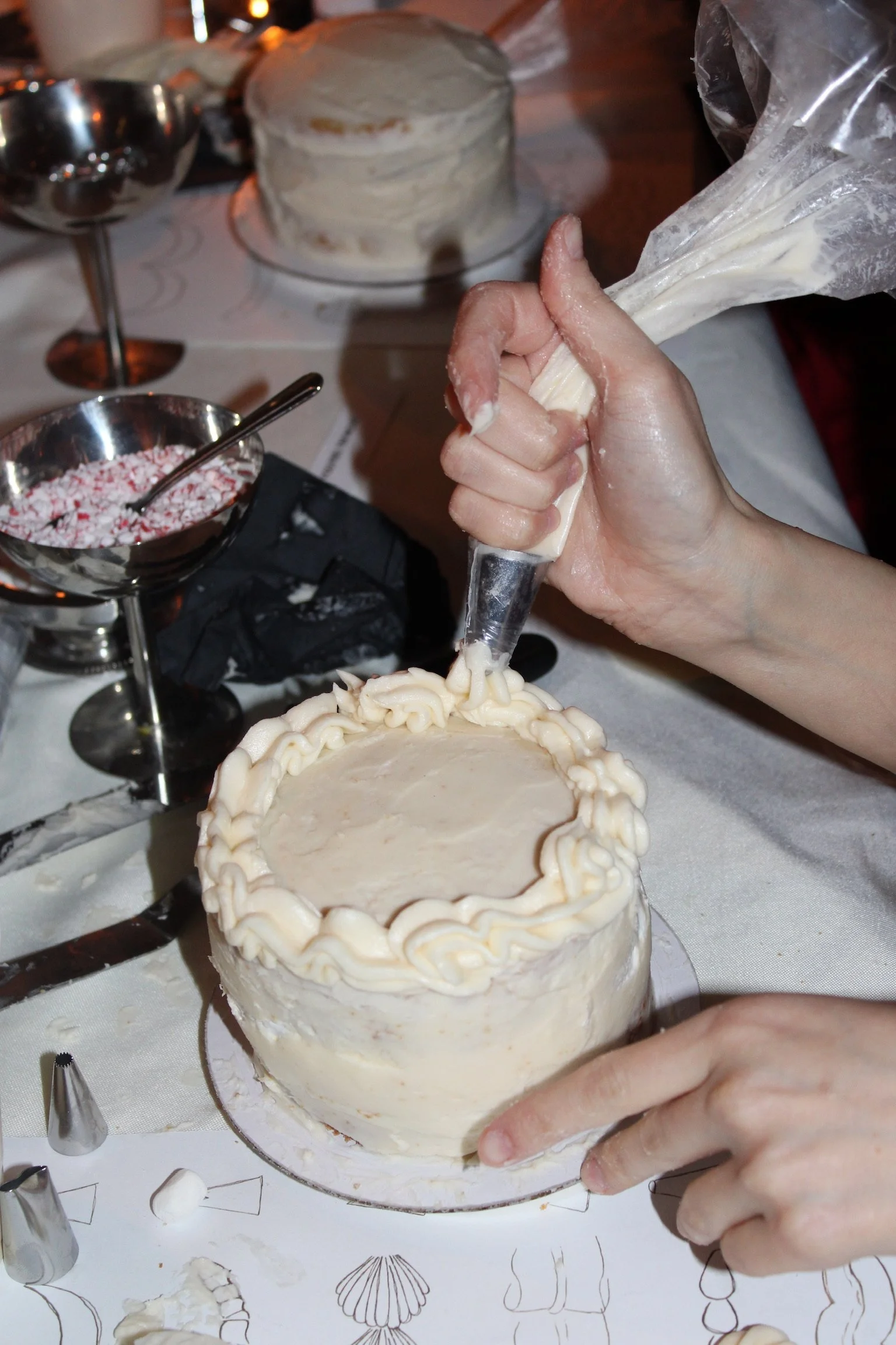 Person decorating a birthday cake with icing, with baking tools and another cake in the background.