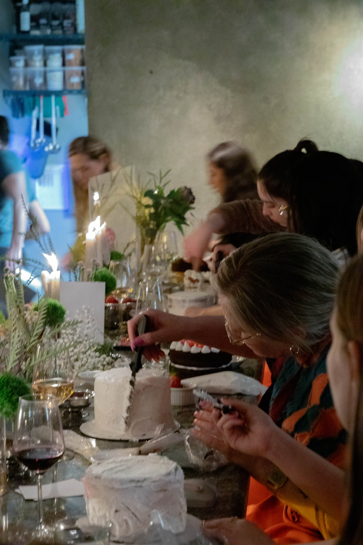 People celebrating at a birthday party, with a decorated table, cakes, candles, flowers, and drinks.
